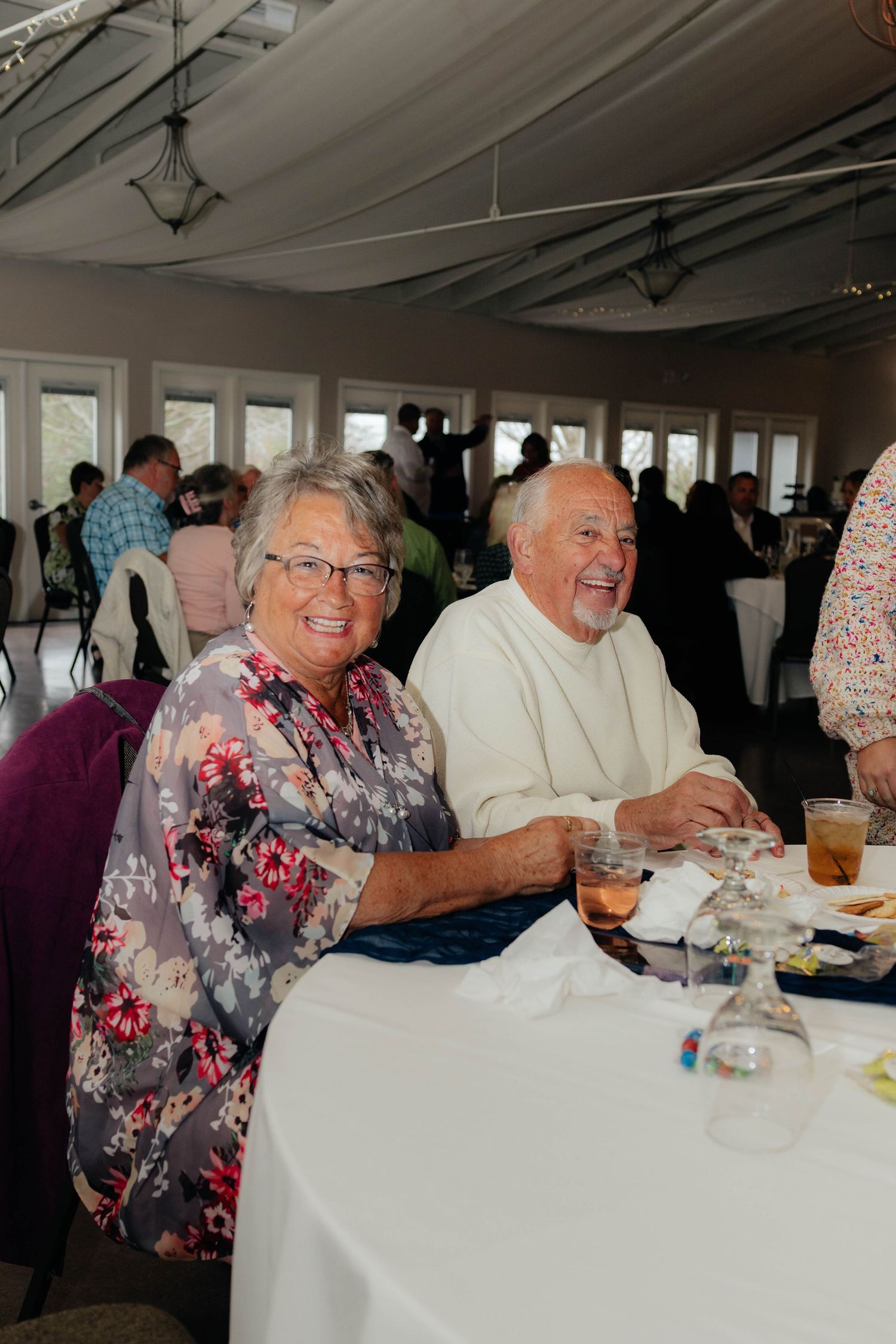 Smiling elderly couple seated together at white-clothed table during wedding reception celebration.