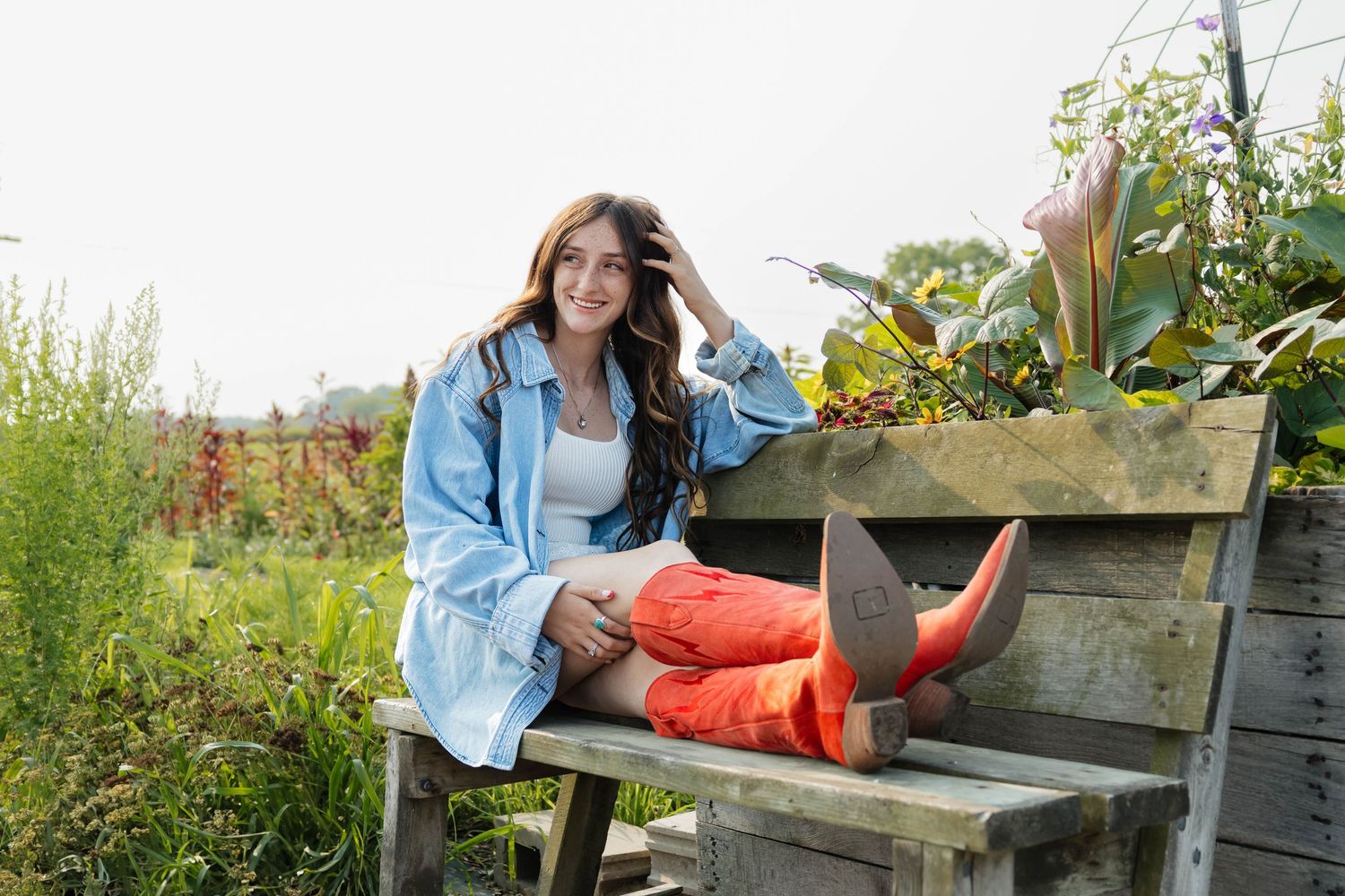 Young woman in denim jacket and red boots relaxing on rustic wooden bench in flower garden.