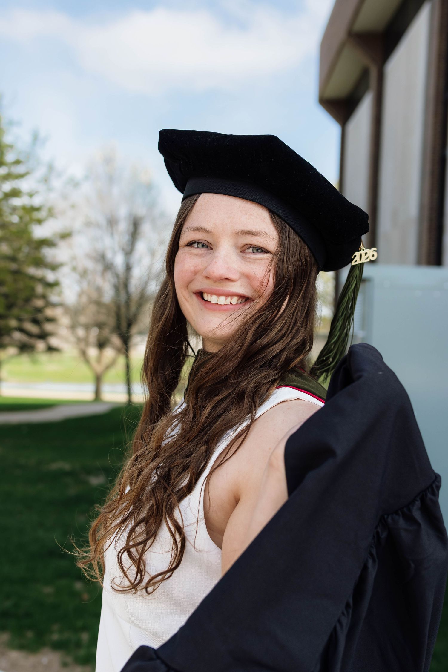 Smiling young woman in doctoral graduation cap and gown posing outdoors on campus in 2025.