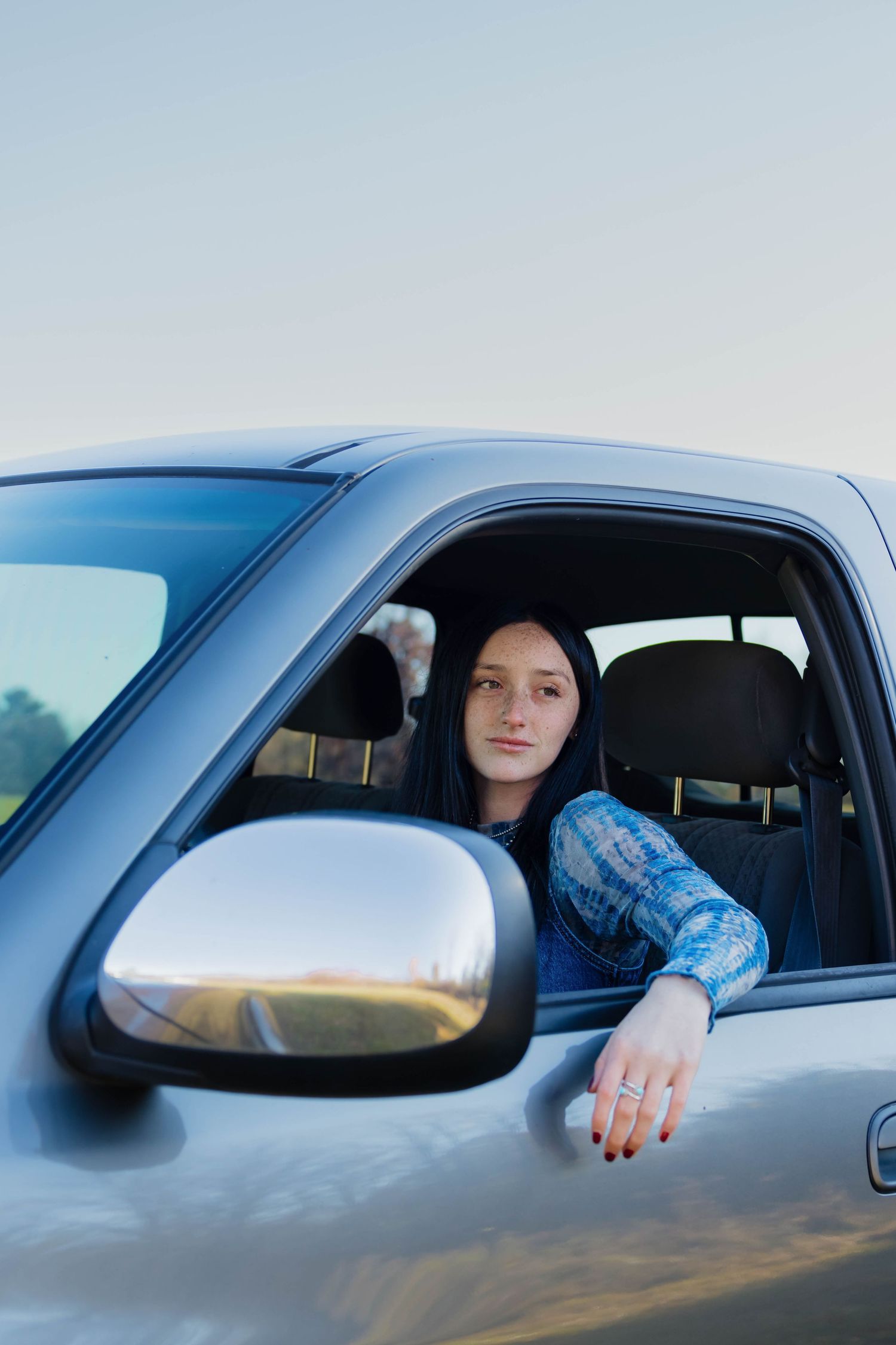 Young woman with dark hair leaning arm out of silver truck window on clear day.