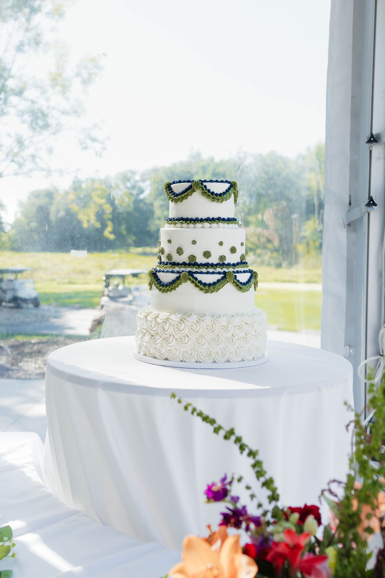 Four-tier white wedding cake decorated with green and navy frosting on white draped table.