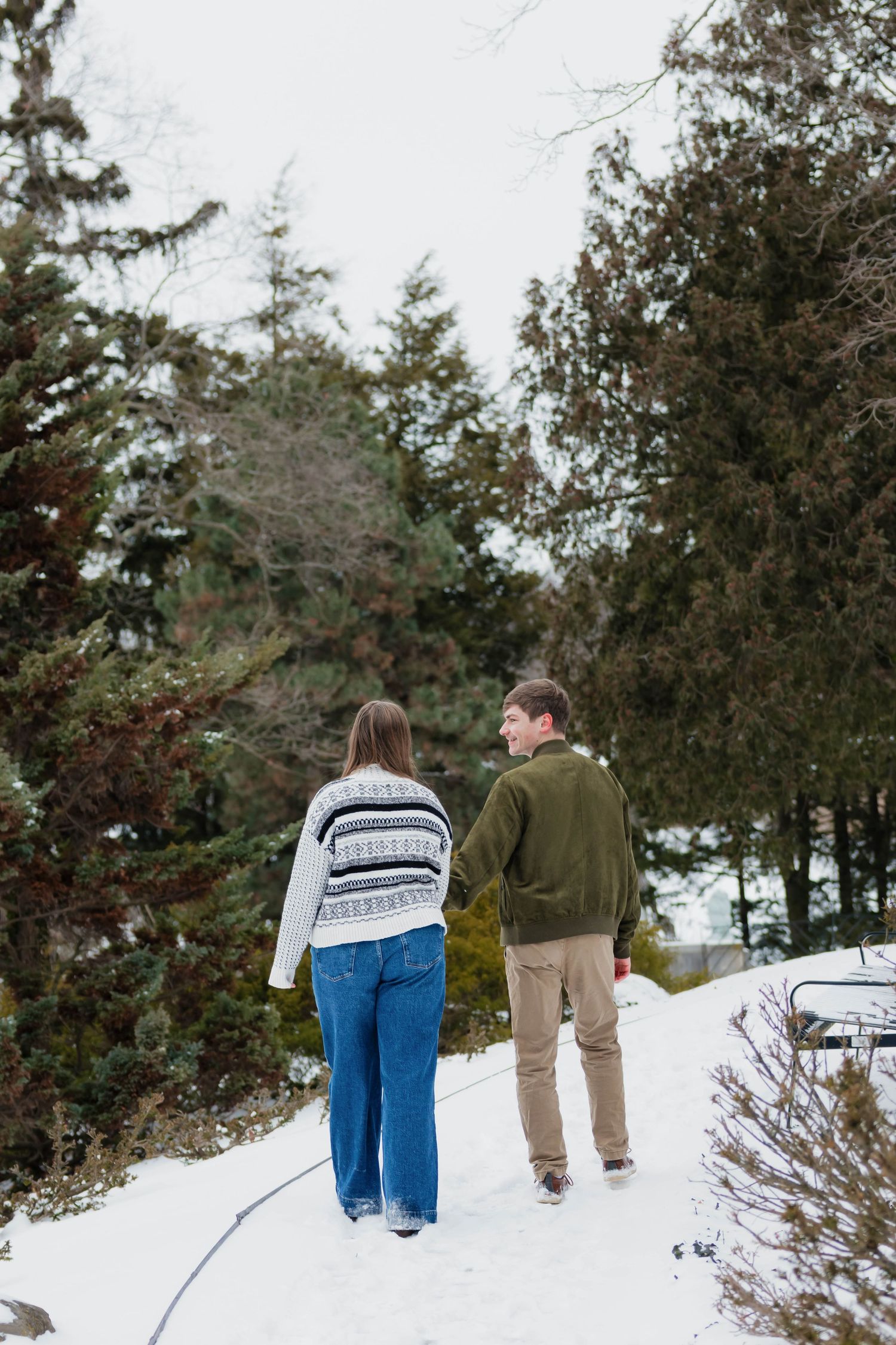 Couple walking hand in hand through a snow-covered path surrounded by evergreen trees.