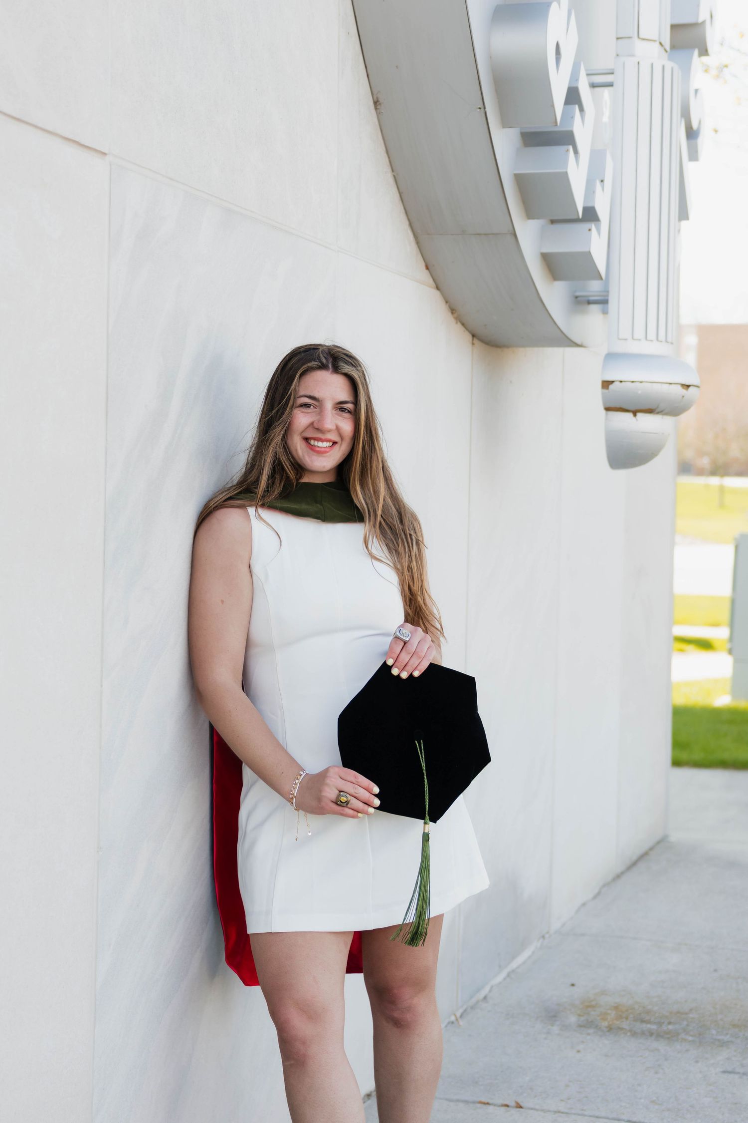 Smiling graduate in white dress holding black mortarboard cap while leaning against white column.