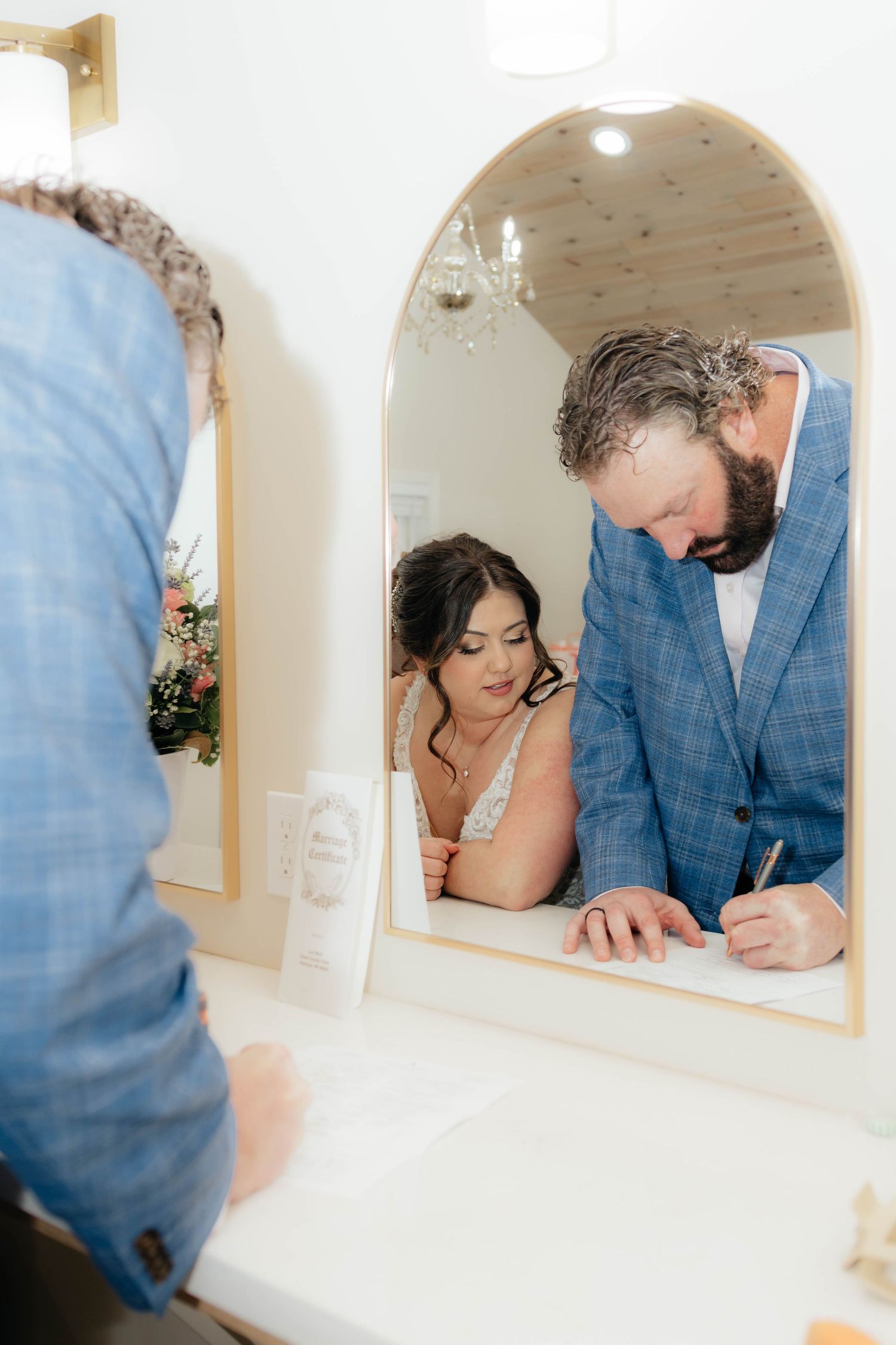 Bride and groom signing wedding certificate reflected in an arched mirror at their wedding venue.