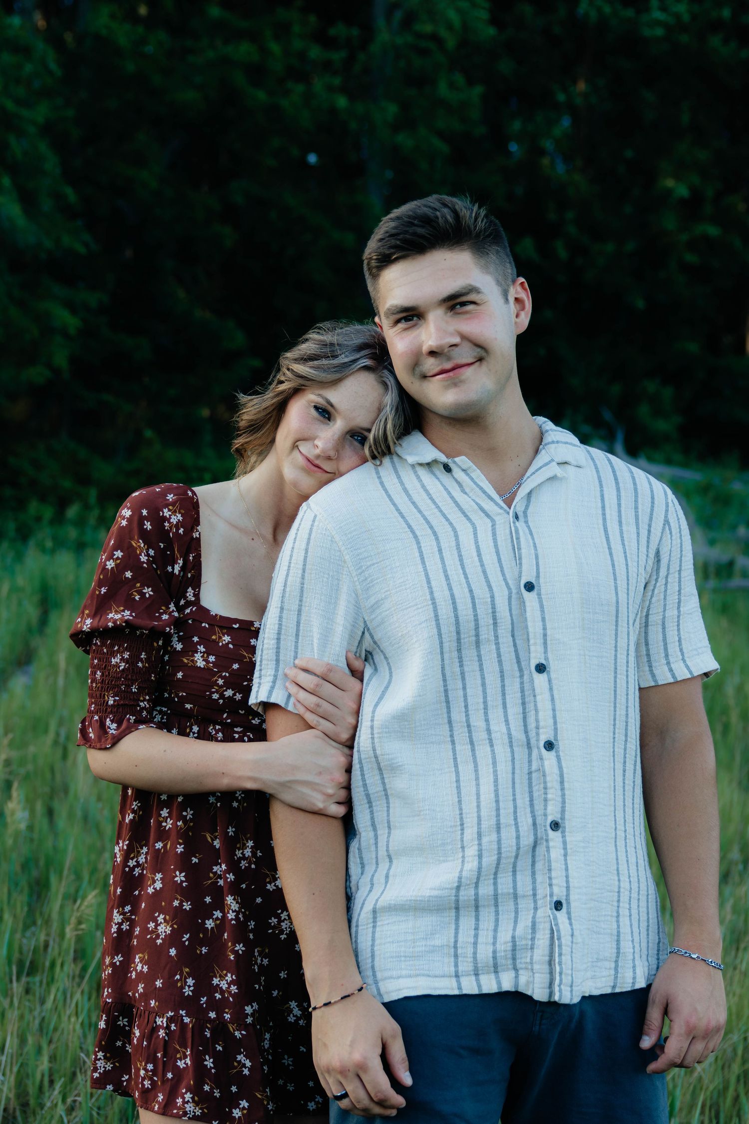 Couple posing in a grassy field at dusk, woman in floral dress leaning on mans shoulder, smiling at camera.