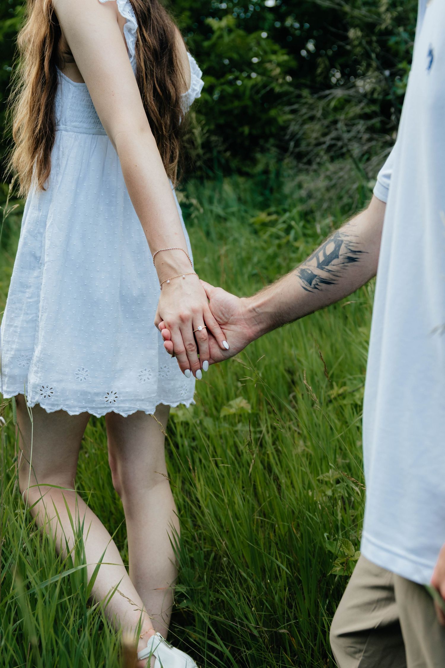 Couple holding hands in lush green field, woman in white eyelet dress, man with floral forearm tattoo.