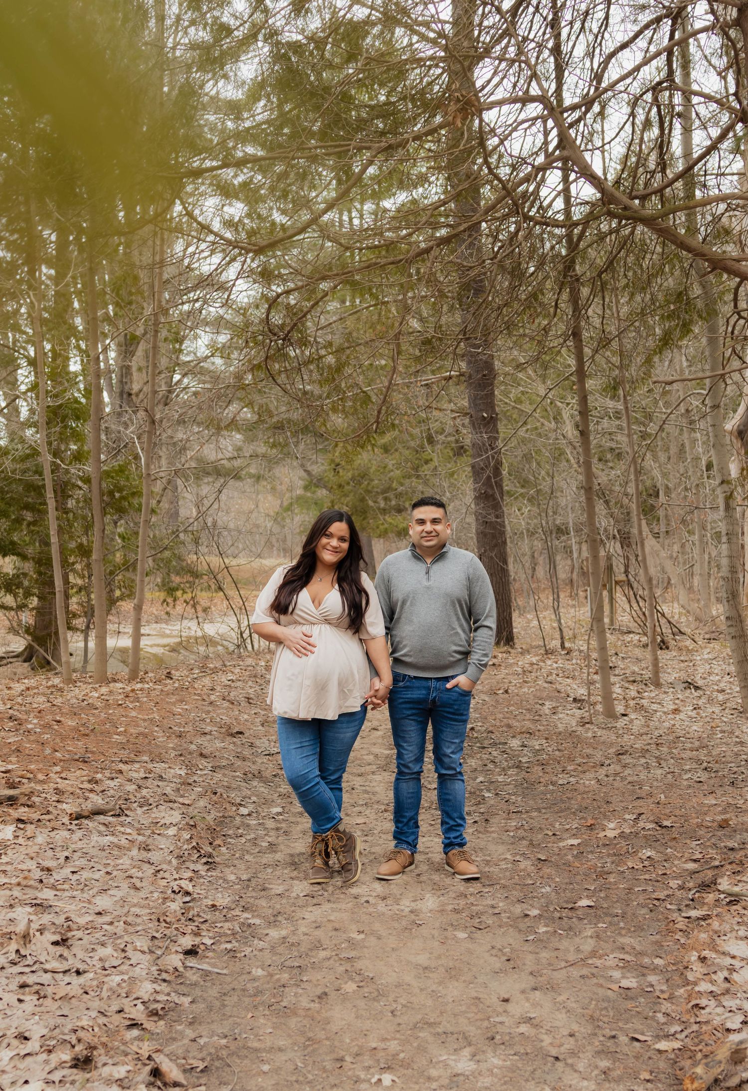 Expecting couple holding hands and smiling while walking on a forest trail during maternity photoshoot.