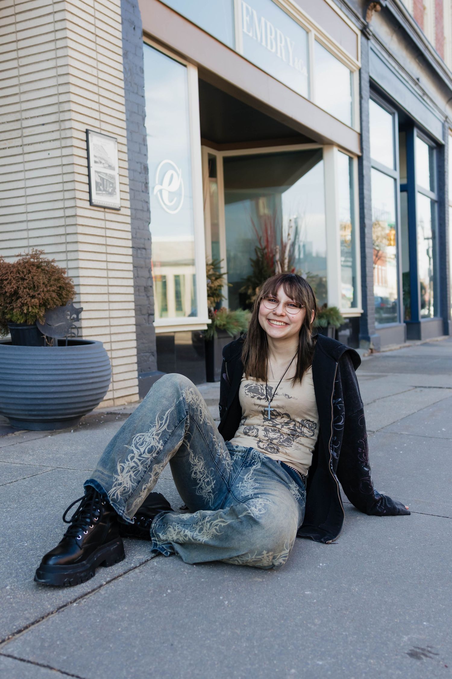 Smiling young woman sitting on sidewalk in front of a store wearing patterned jeans and black jacket.