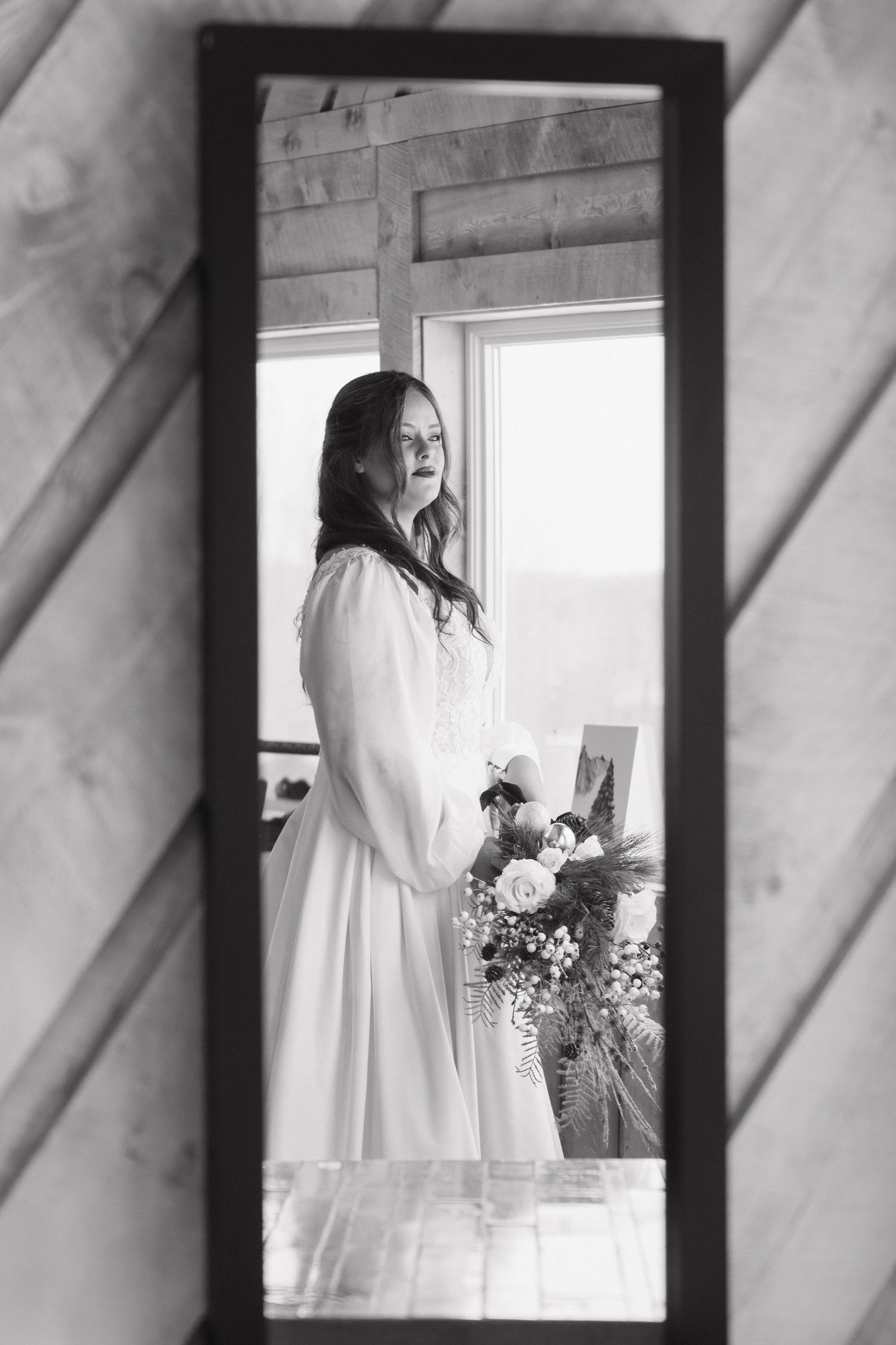 Black and white mirror reflection of bride in long-sleeve wedding gown holding a floral bouquet.