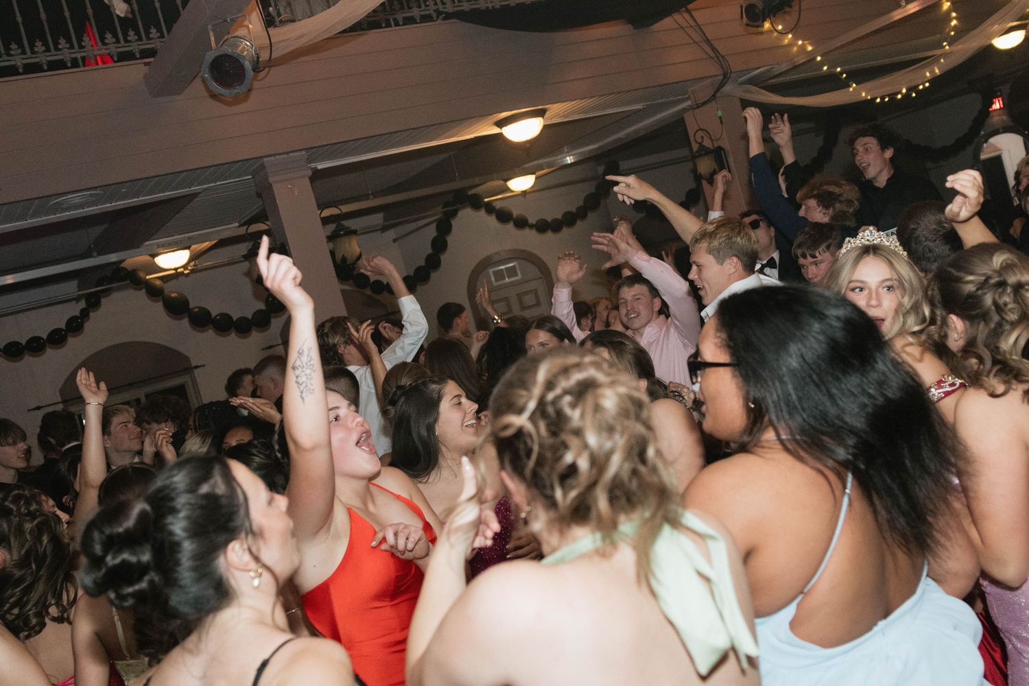 A lively crowd of young people dancing and celebrating at a formal party or prom event indoors.