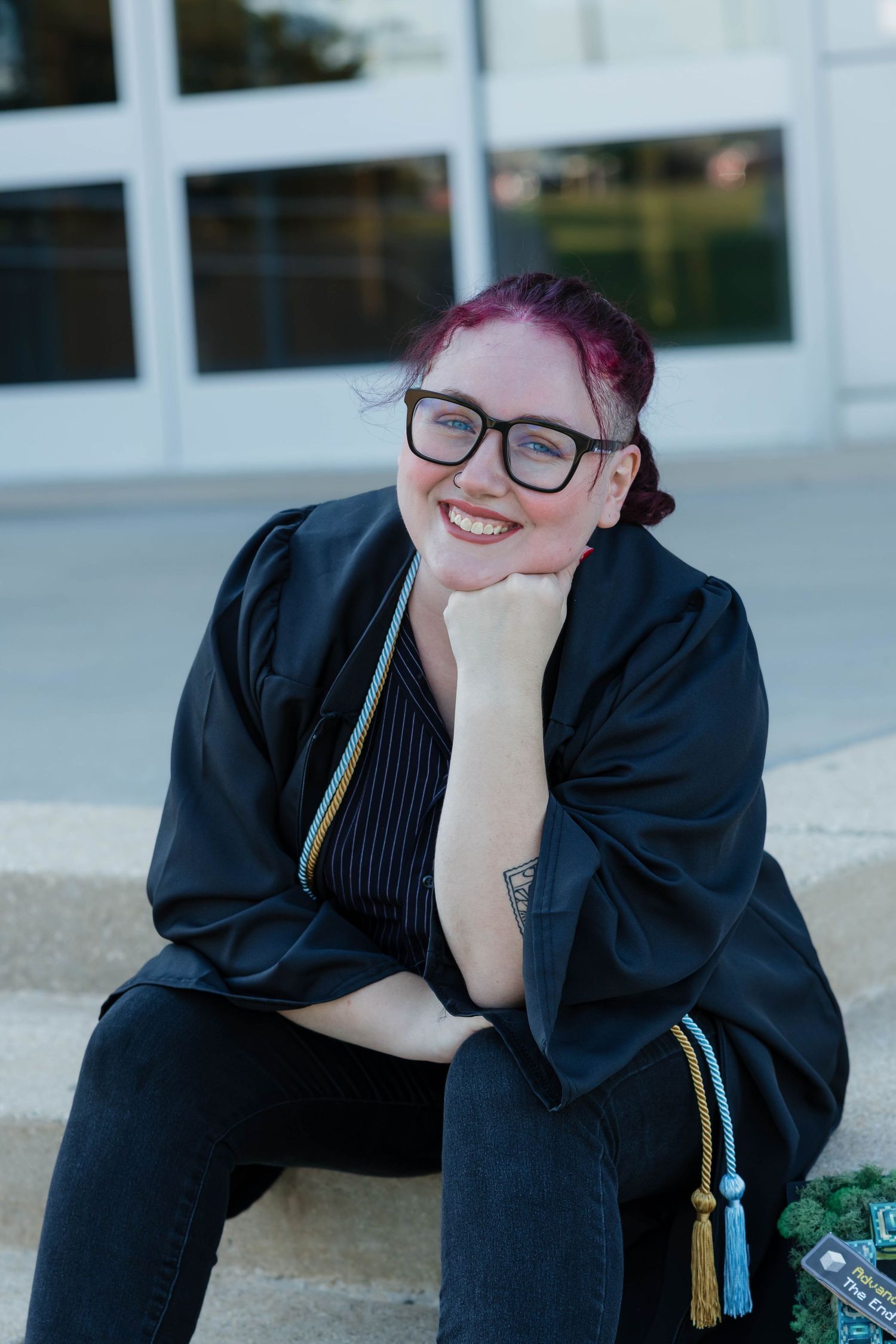 Happy graduate in black gown sitting on steps with colorful tassels smiling at camera.