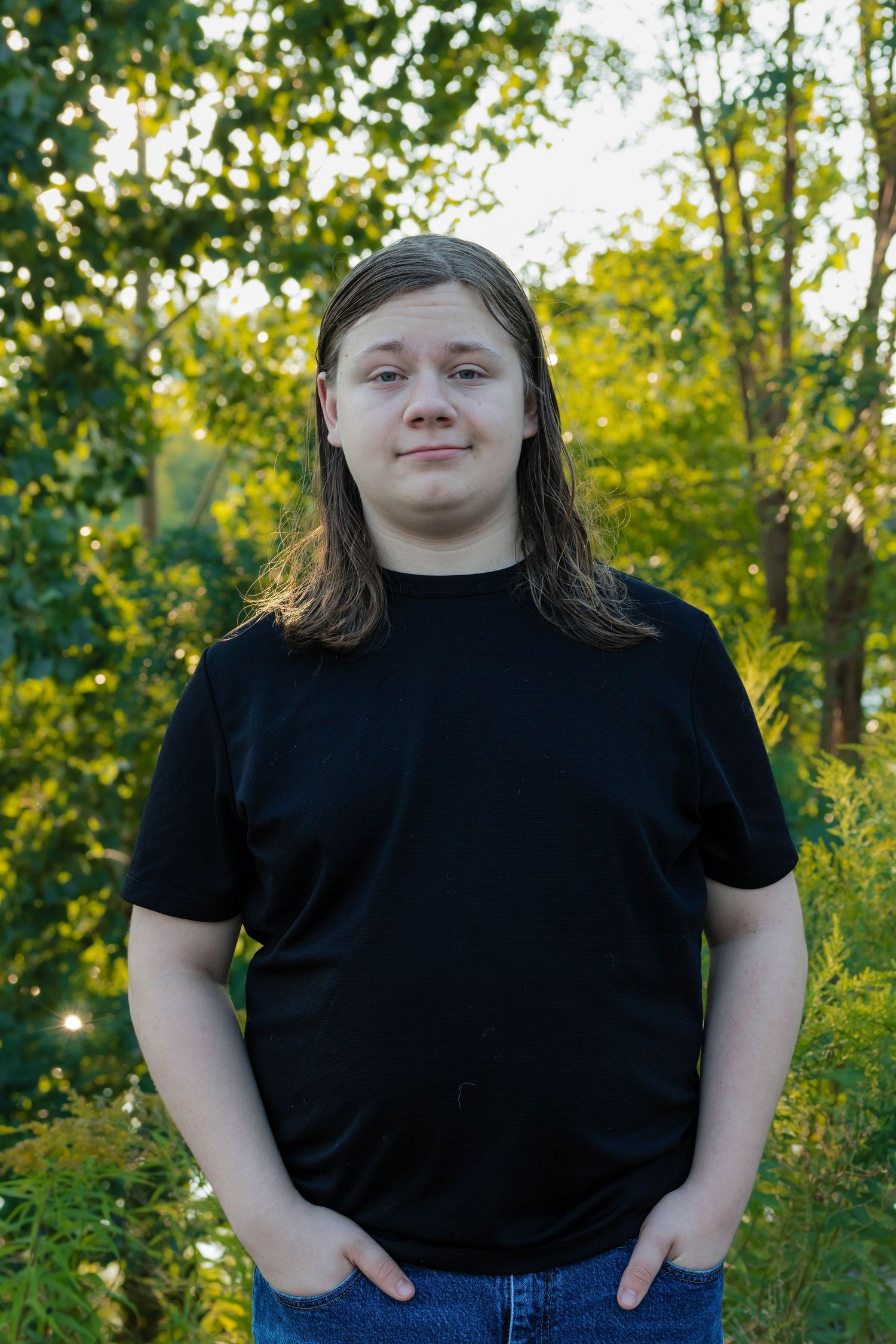 A teenage boy with long hair wearing a black t-shirt stands smiling in a sunlit green outdoor setting.