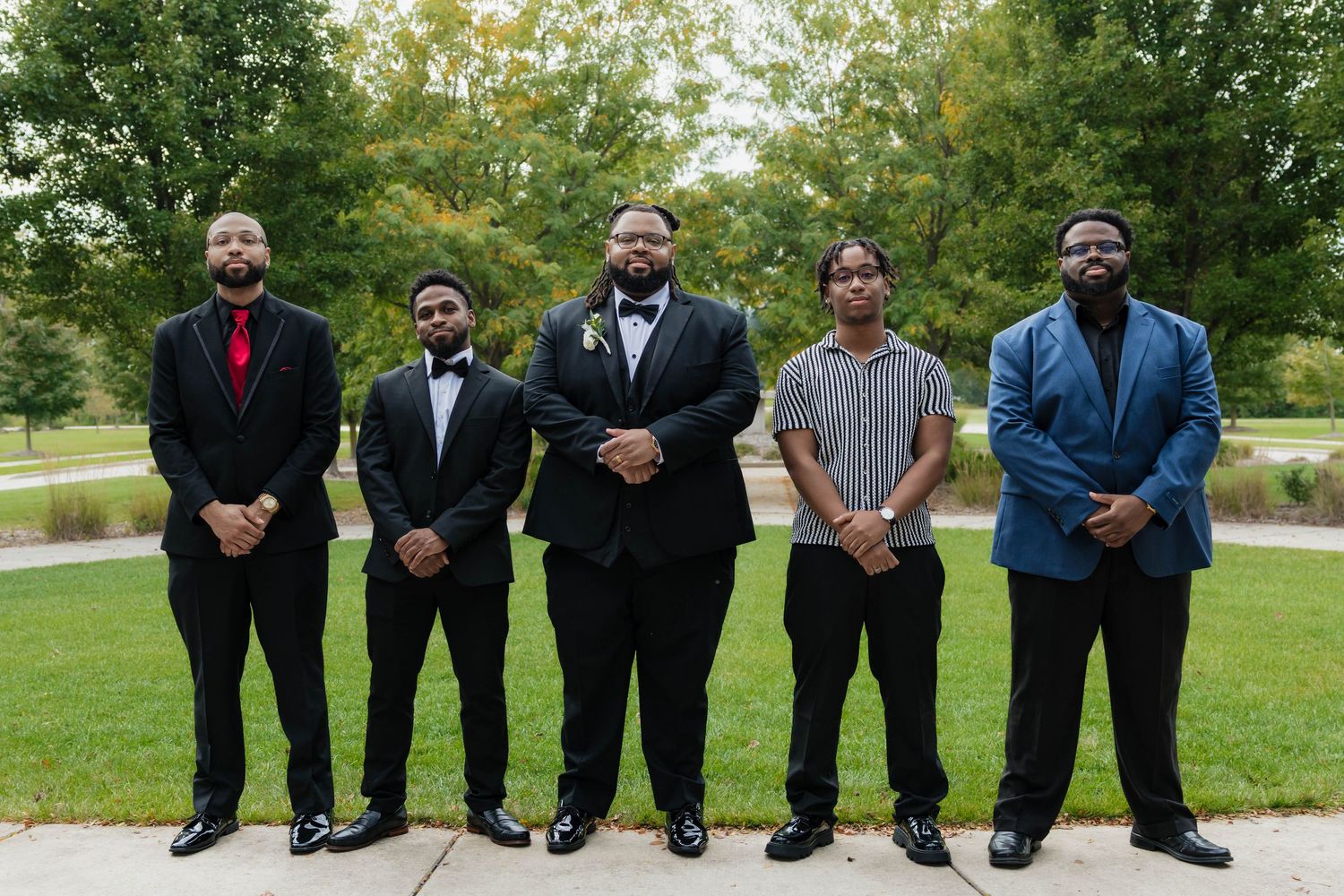 Groom in tuxedo stands with four groomsmen in formal attire outdoors at wedding.