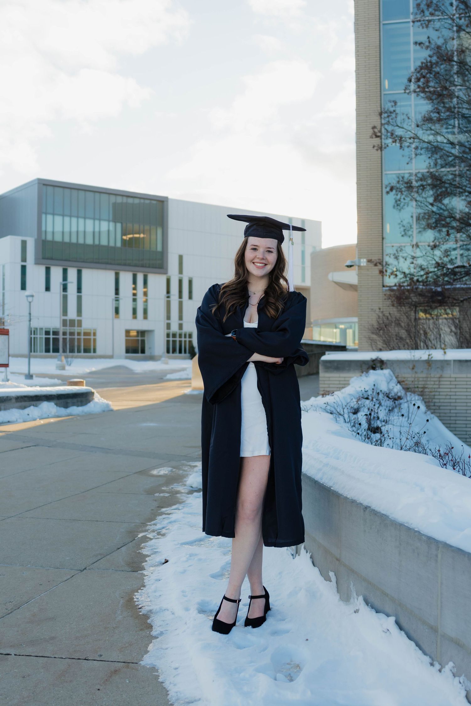 Brunette graduate in cap and gown posing confidently with arms crossed on a snowy campus.