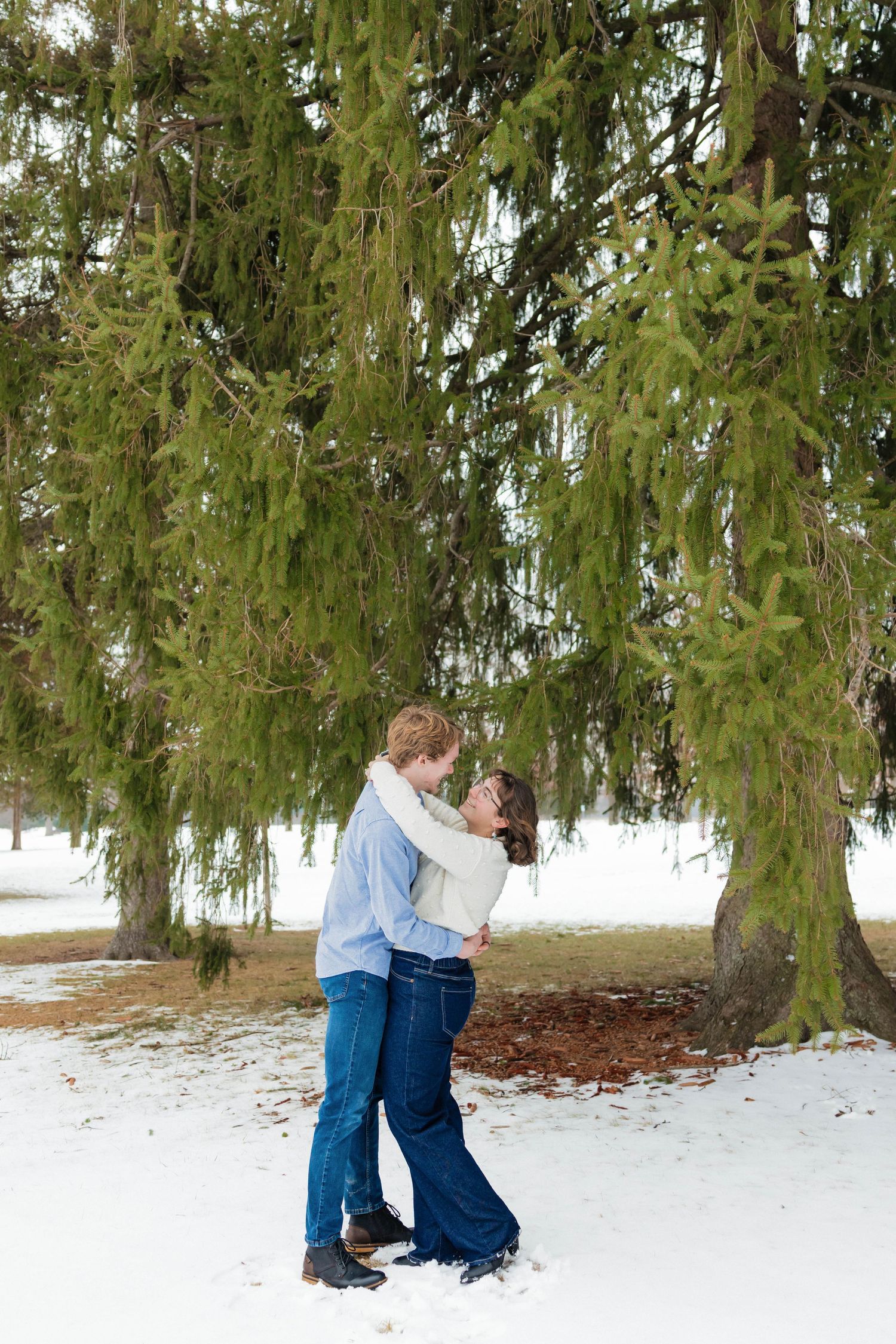 Couple sharing a romantic embrace under snow-dusted evergreen trees in a winter park.