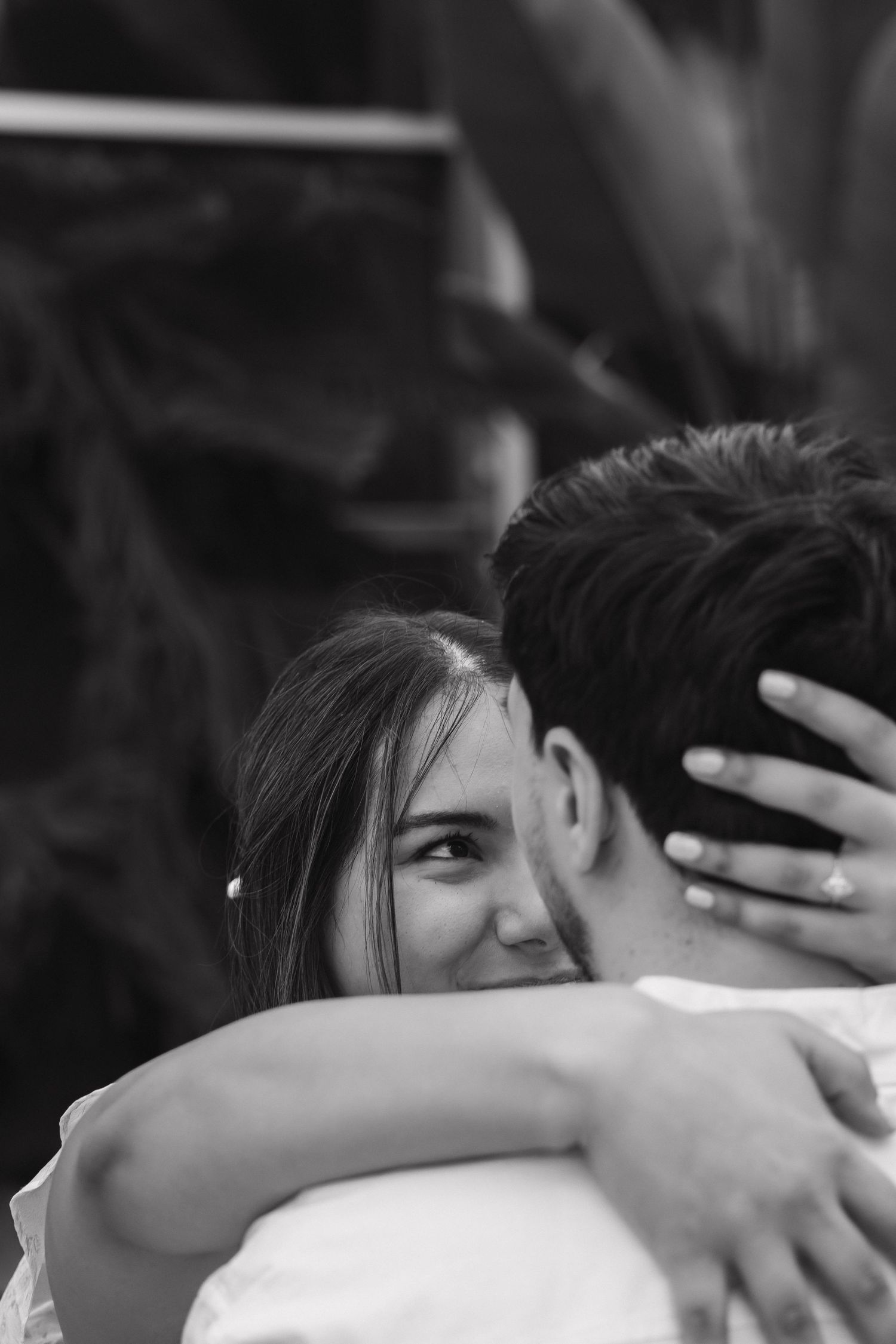 Black and white photo of a woman smiling while embracing her partner during engagement shoot.