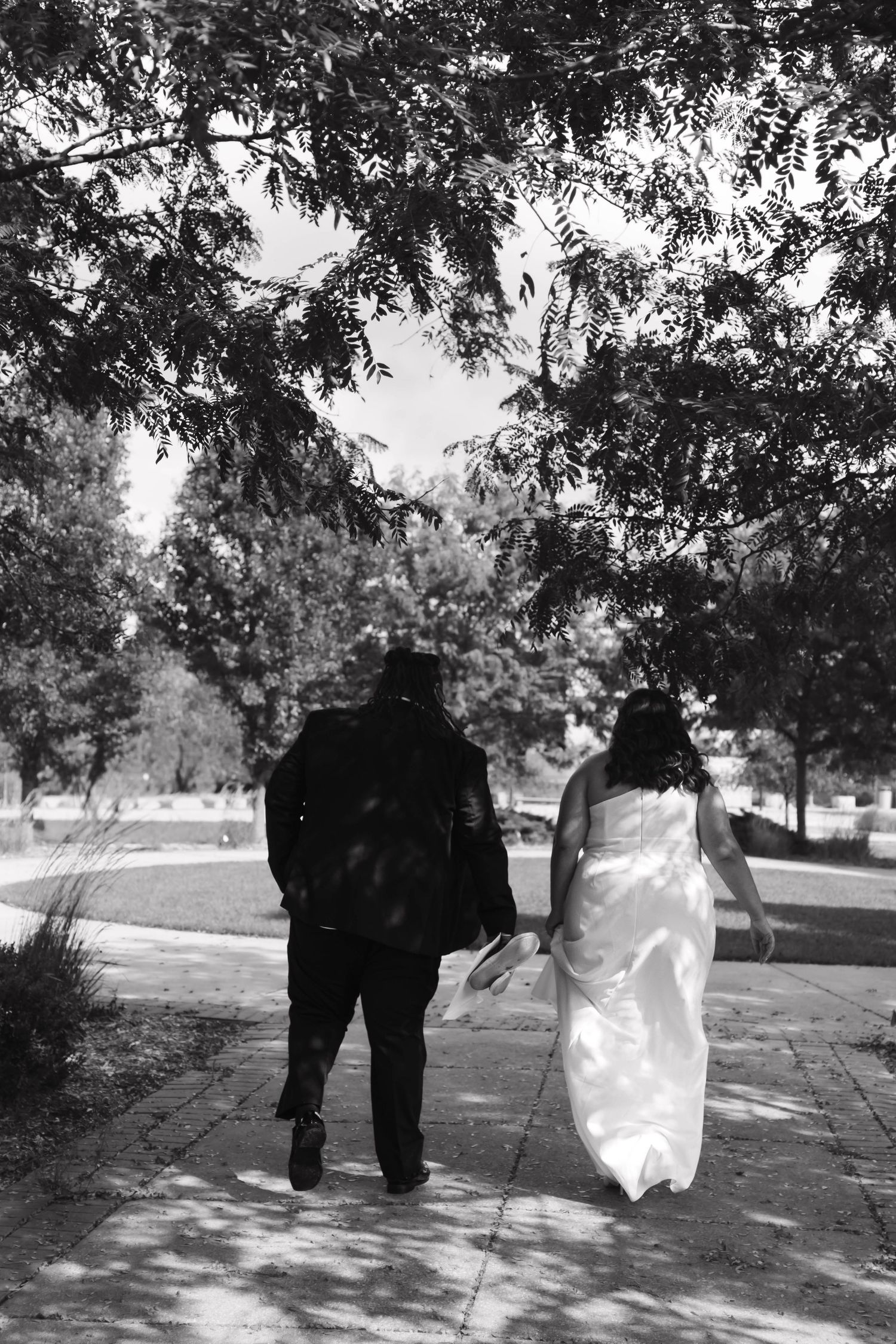 Black and white photo of a bride and groom walking together along a park path.