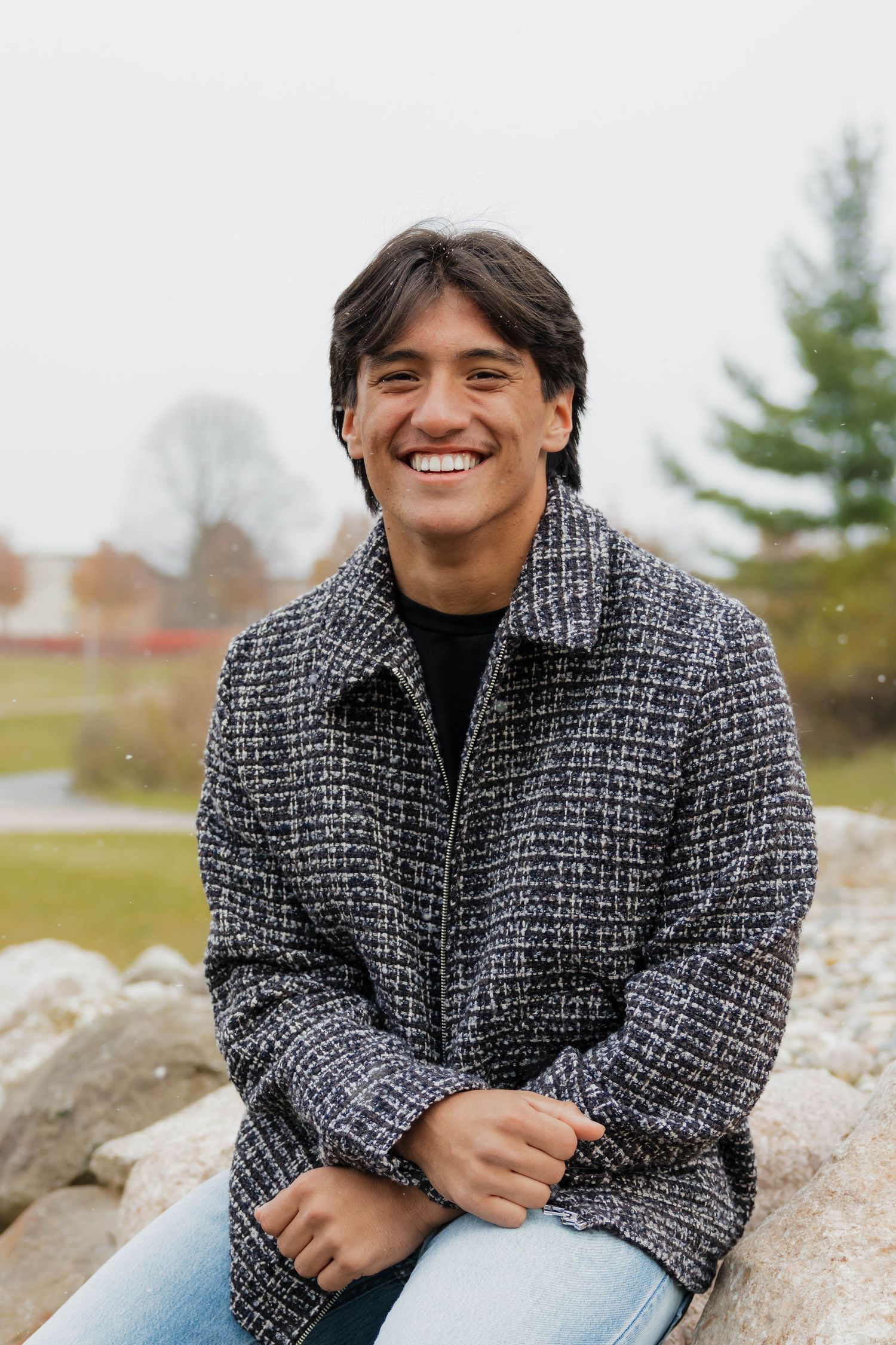 Smiling young man in tweed jacket sitting on rocks outdoors during overcast autumn day.