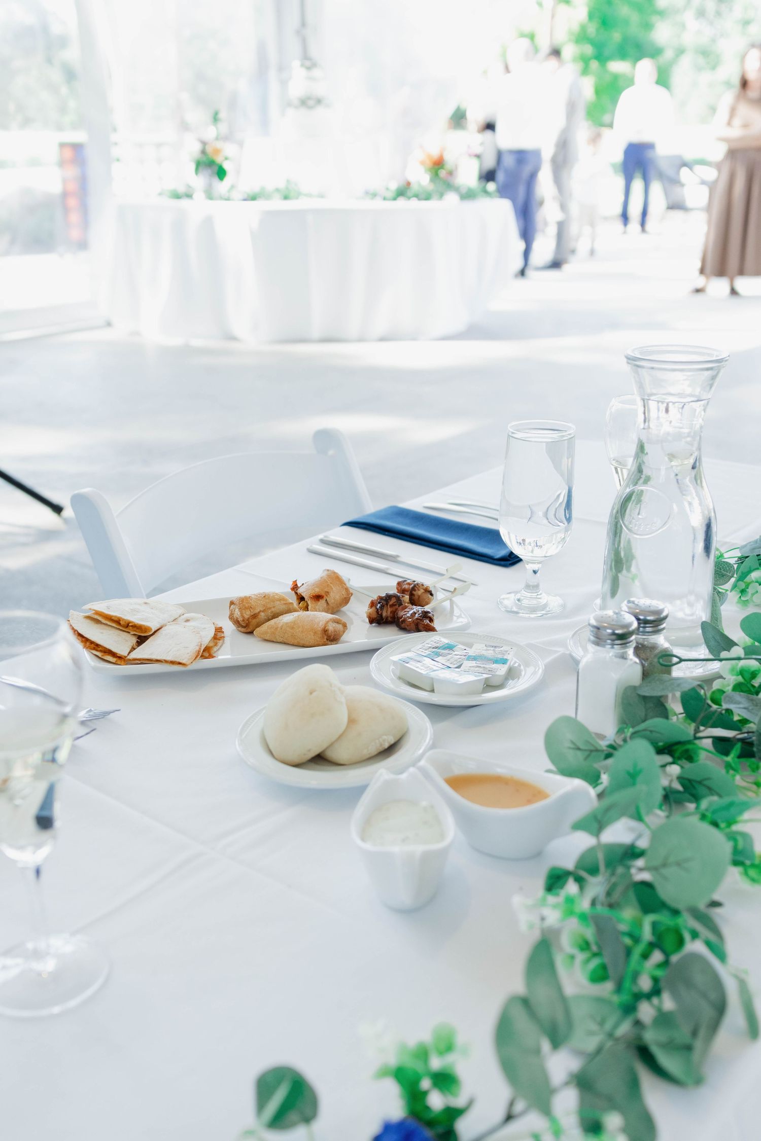 Elegant white wedding reception table with appetizers, bread rolls, glassware, and green eucalyptus garland centerpiece.