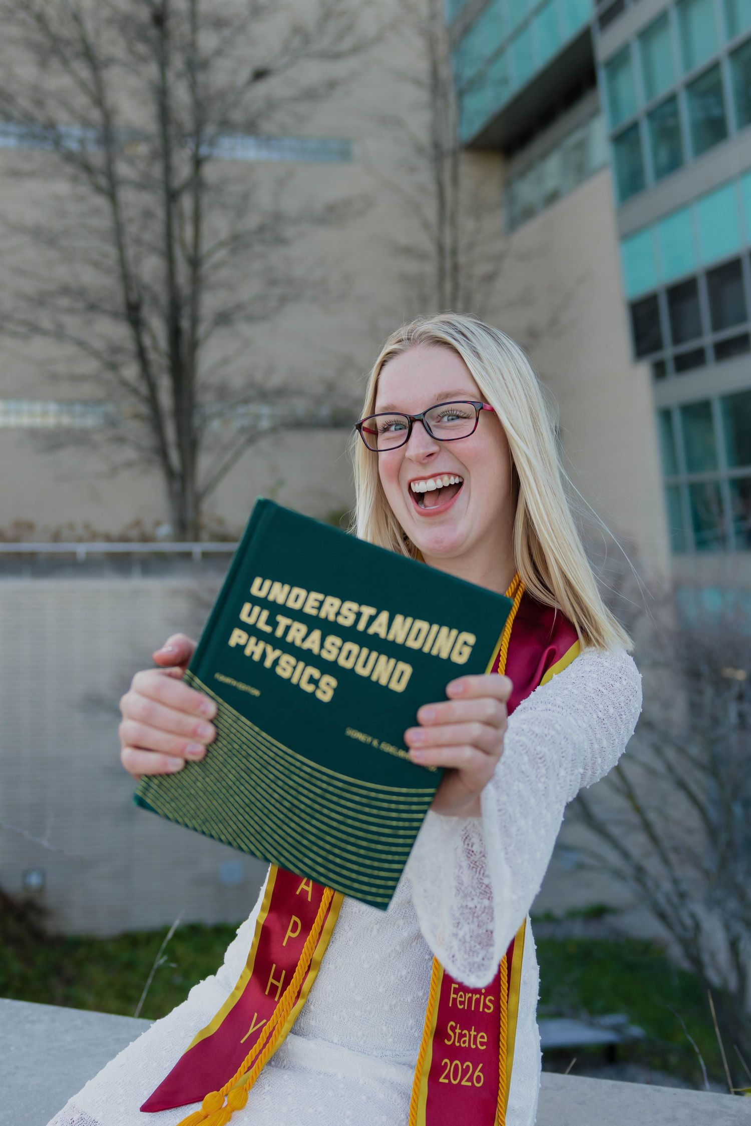 Laughing female Ferris State 2026 graduate holds Understanding Ultrasound Physics textbook outdoors on campus.