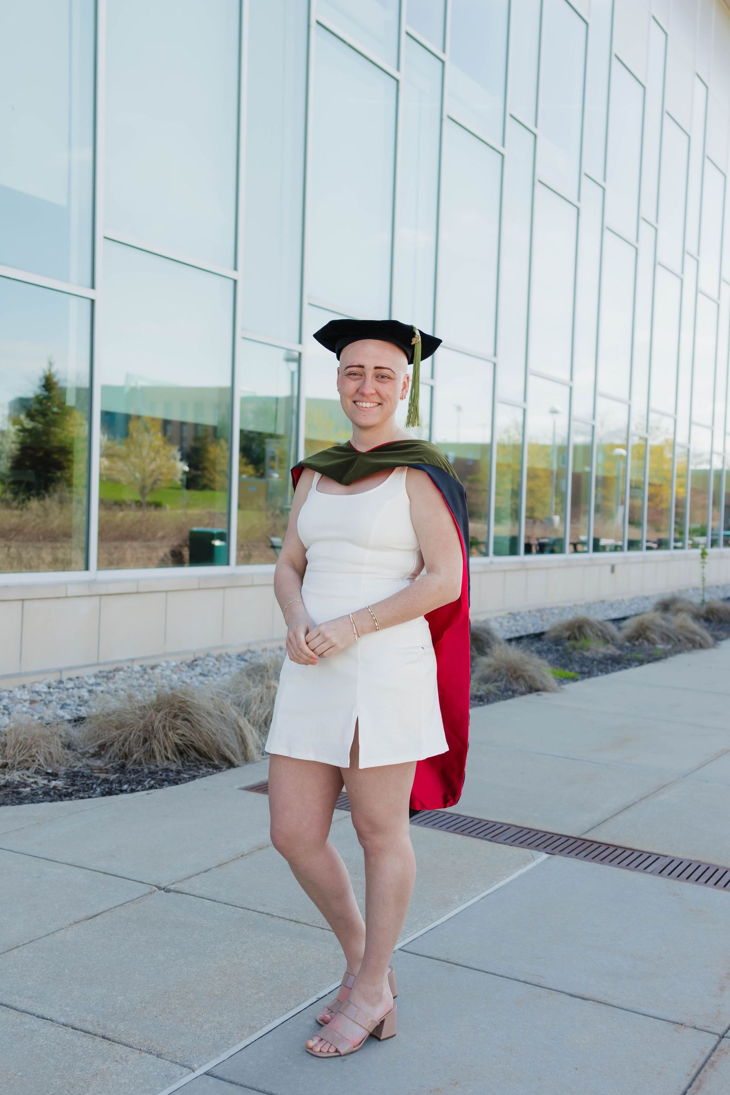 Smiling graduate in white outfit wearing doctoral cap with green and red hood outside university.