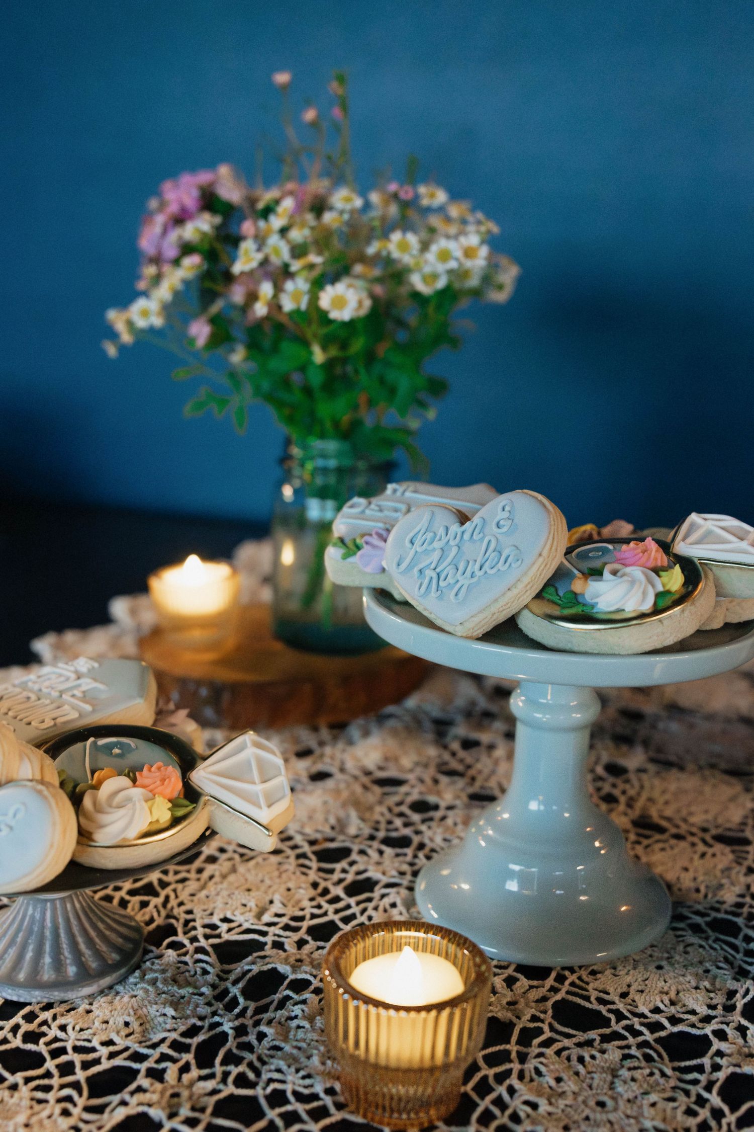 Romantic wedding dessert table with heart-shaped cookies on a white cake stand, candles, and wildflower bouquet.