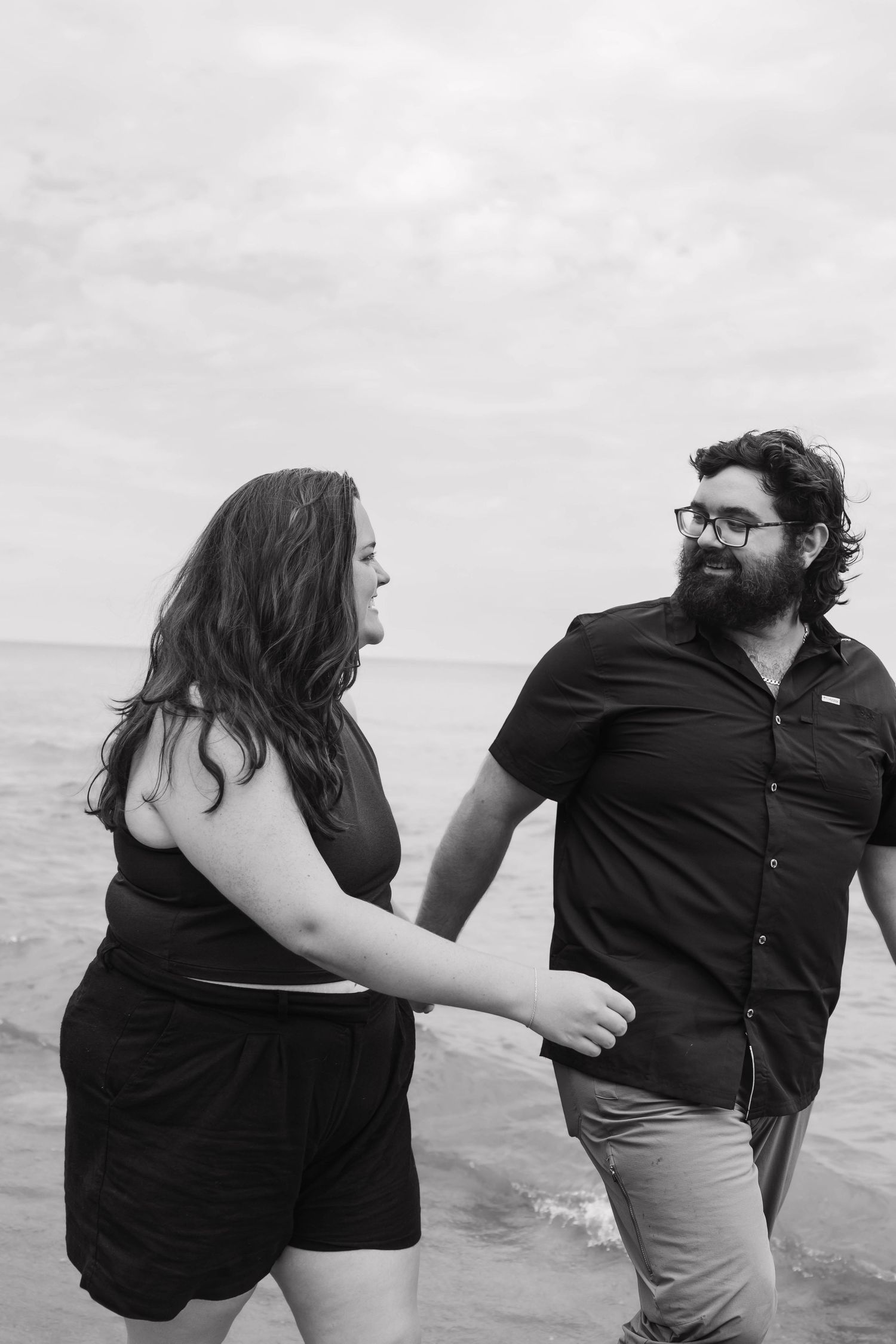 A black and white photo of a couple holding hands and smiling at each other by the ocean.