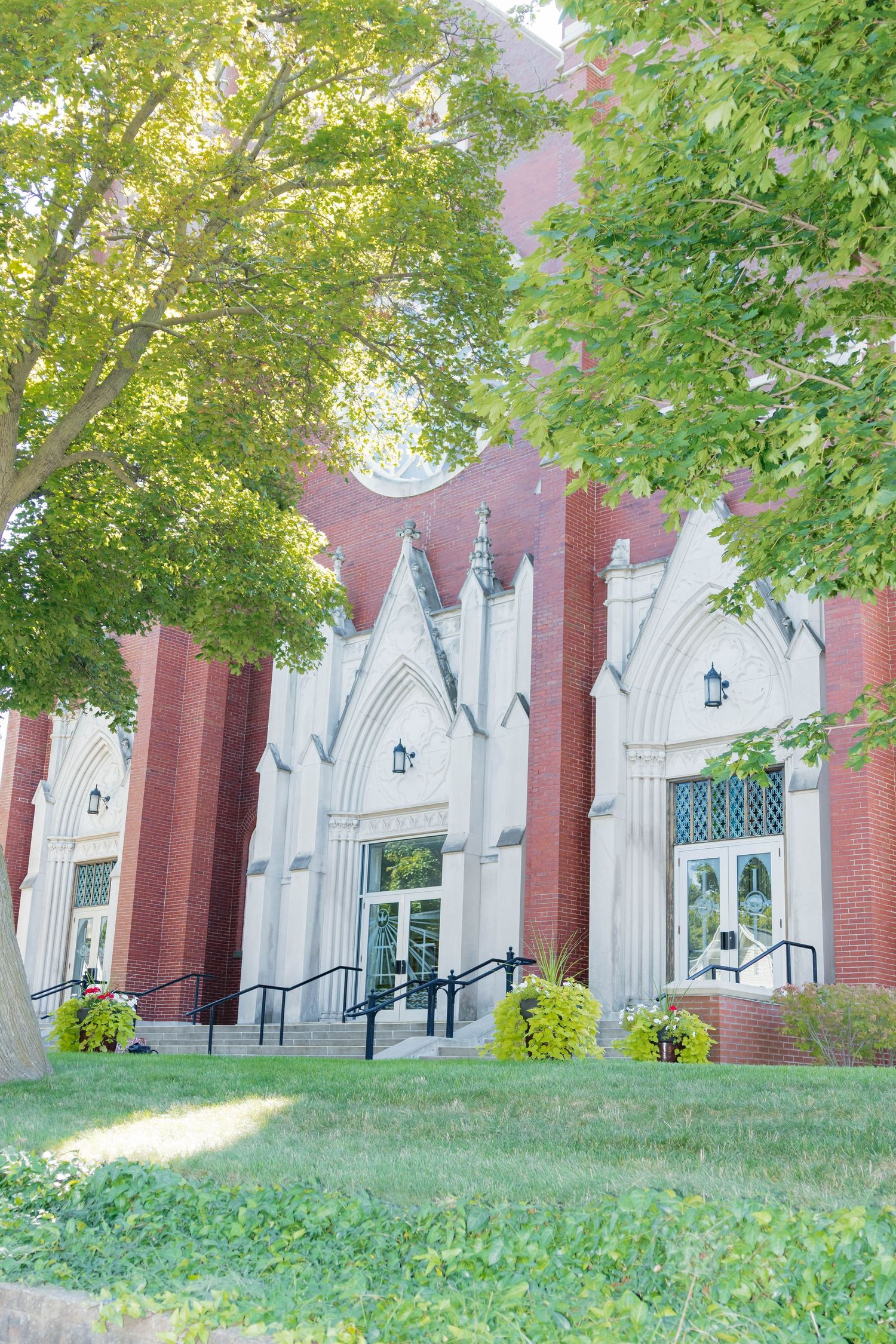 Historic red brick church with Gothic white stone arched entrance surrounded by lush green trees.