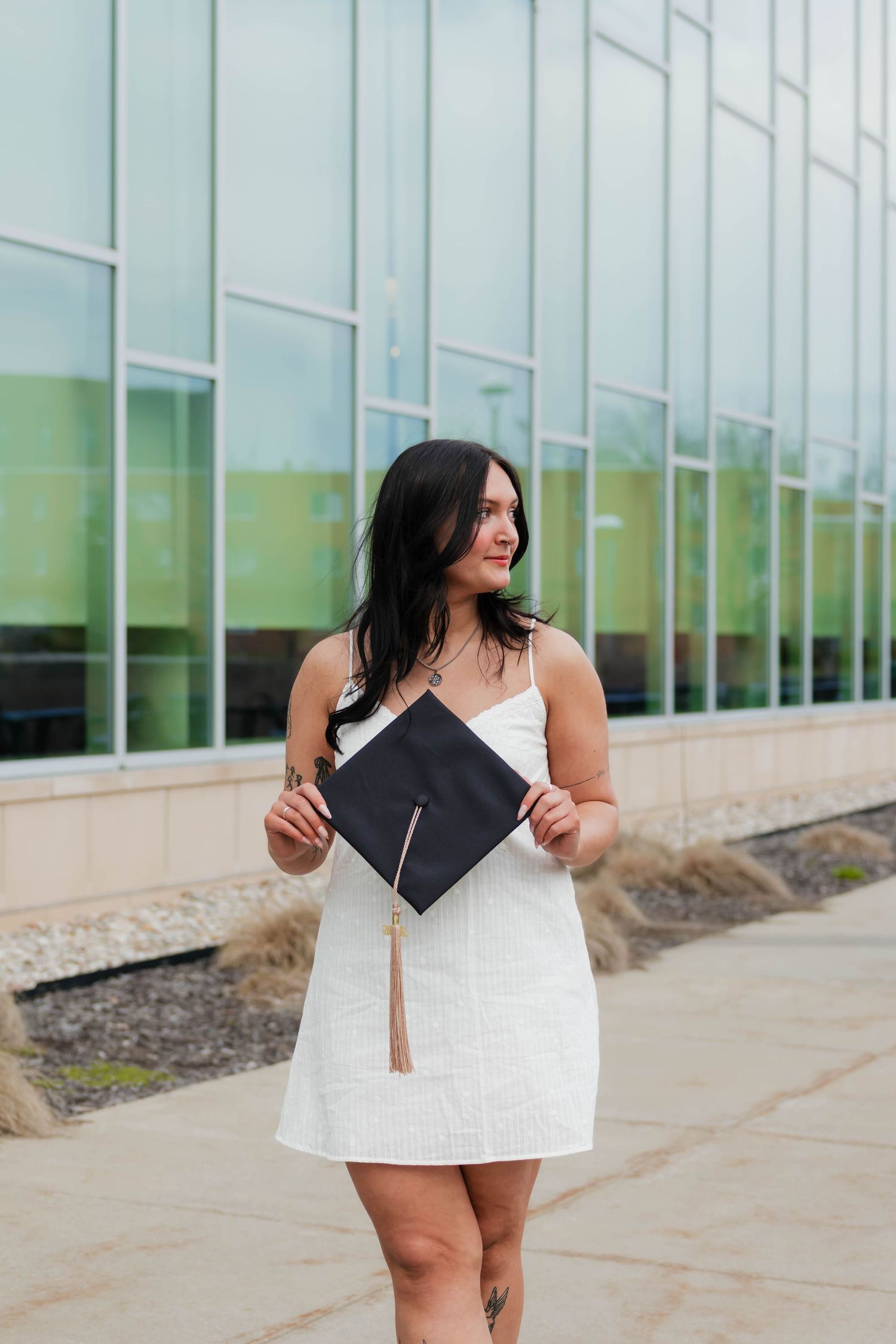 Smiling brunette woman in white dress holding black graduation cap outside glass building.