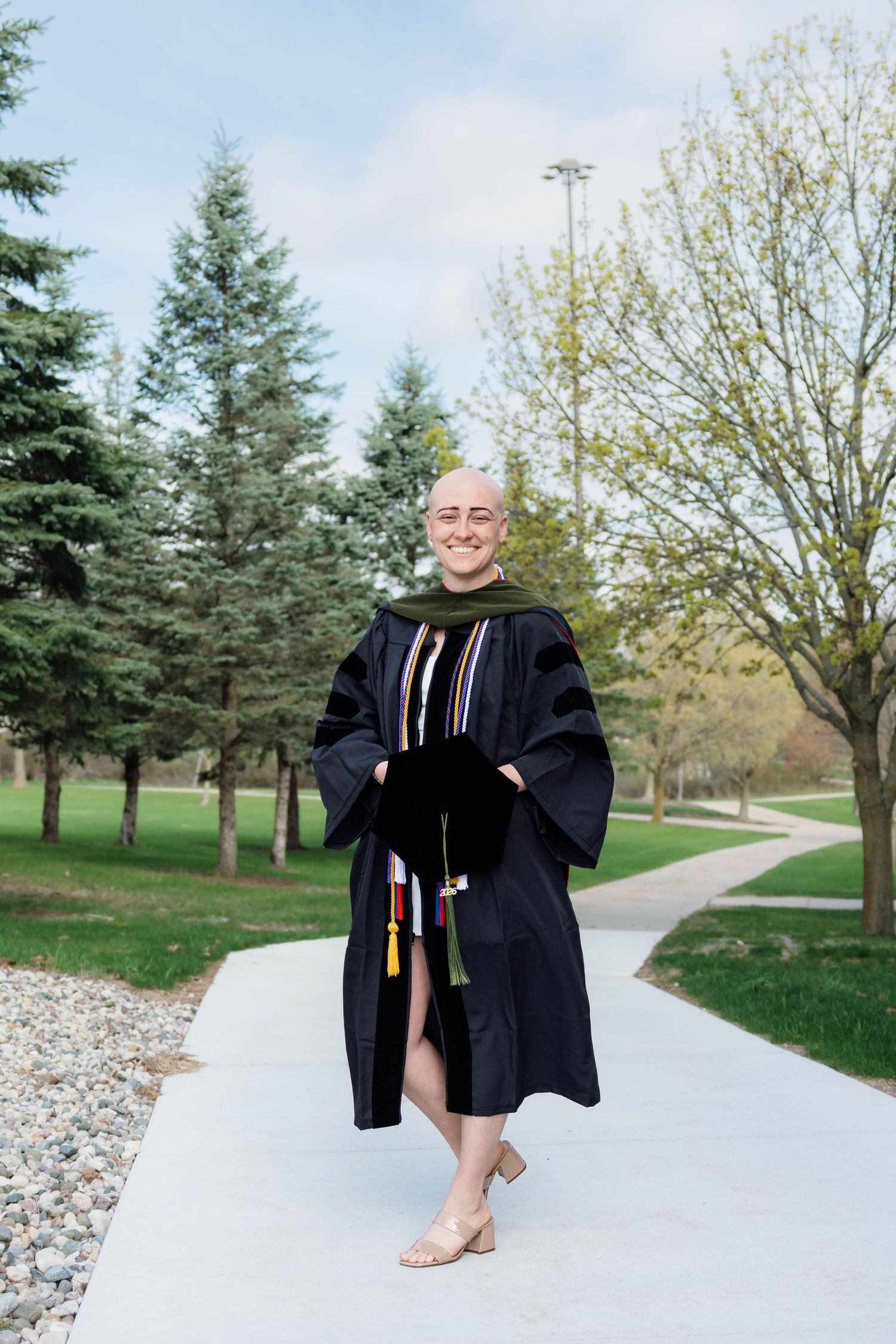 Bald graduate in doctoral regalia with colorful hood and tassels smiles on campus pathway.