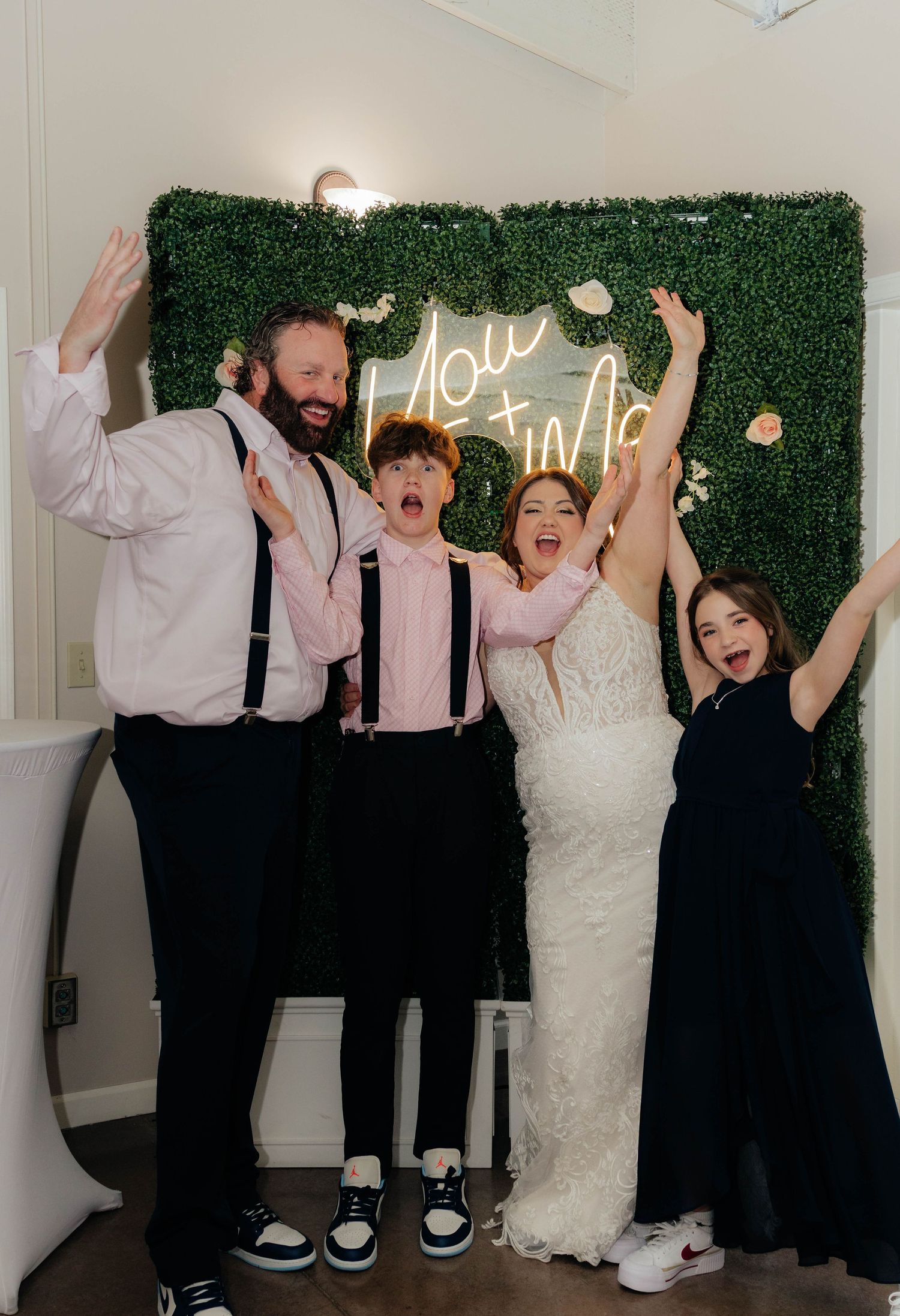 Family of four celebrates joyfully in front of green hedge wall with glowing neon wedding sign.