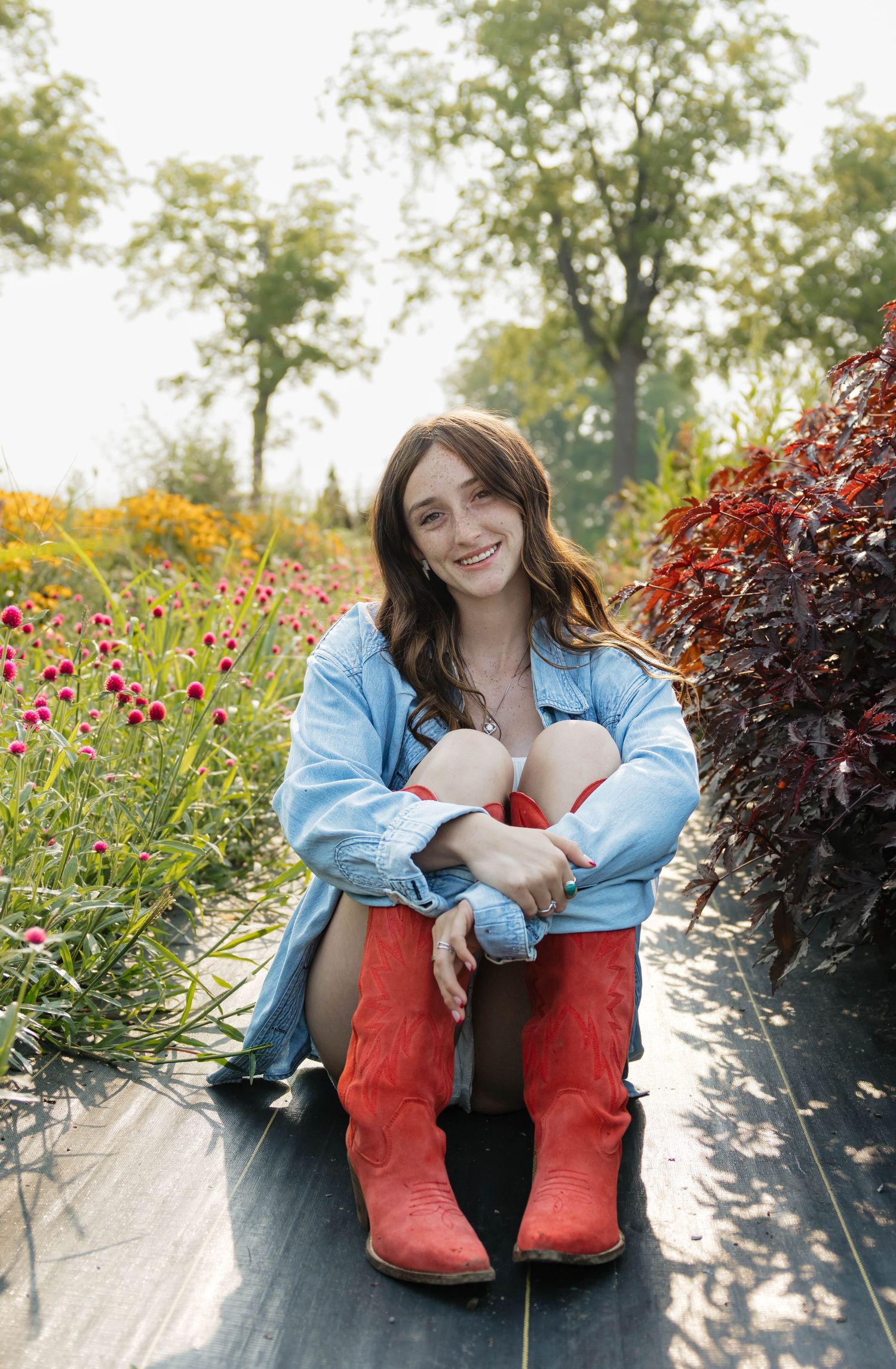 Smiling girl in denim shirt and red cowboy boots sitting on path among colorful garden flowers.