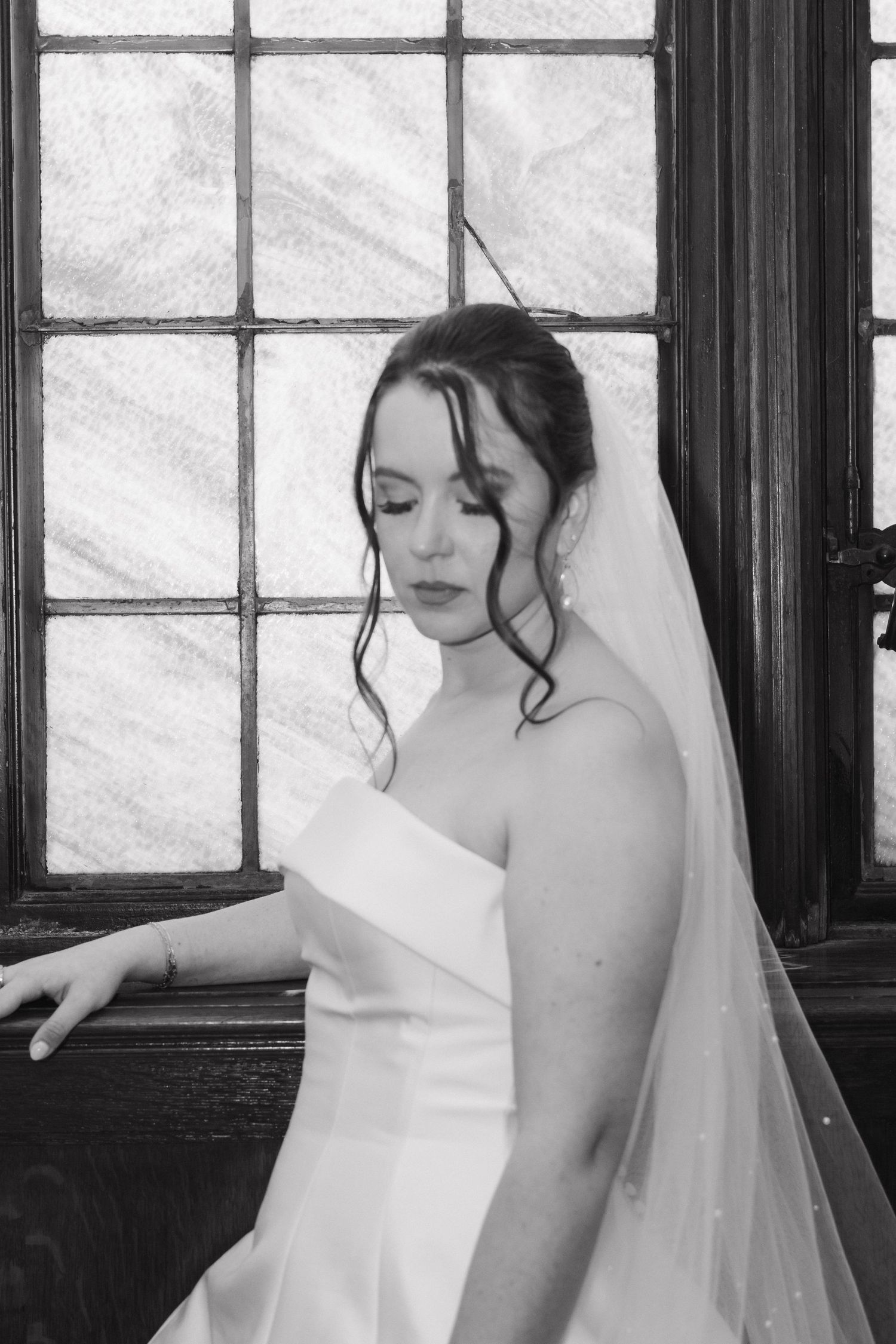Black and white portrait of bride in strapless gown and veil by vintage window.