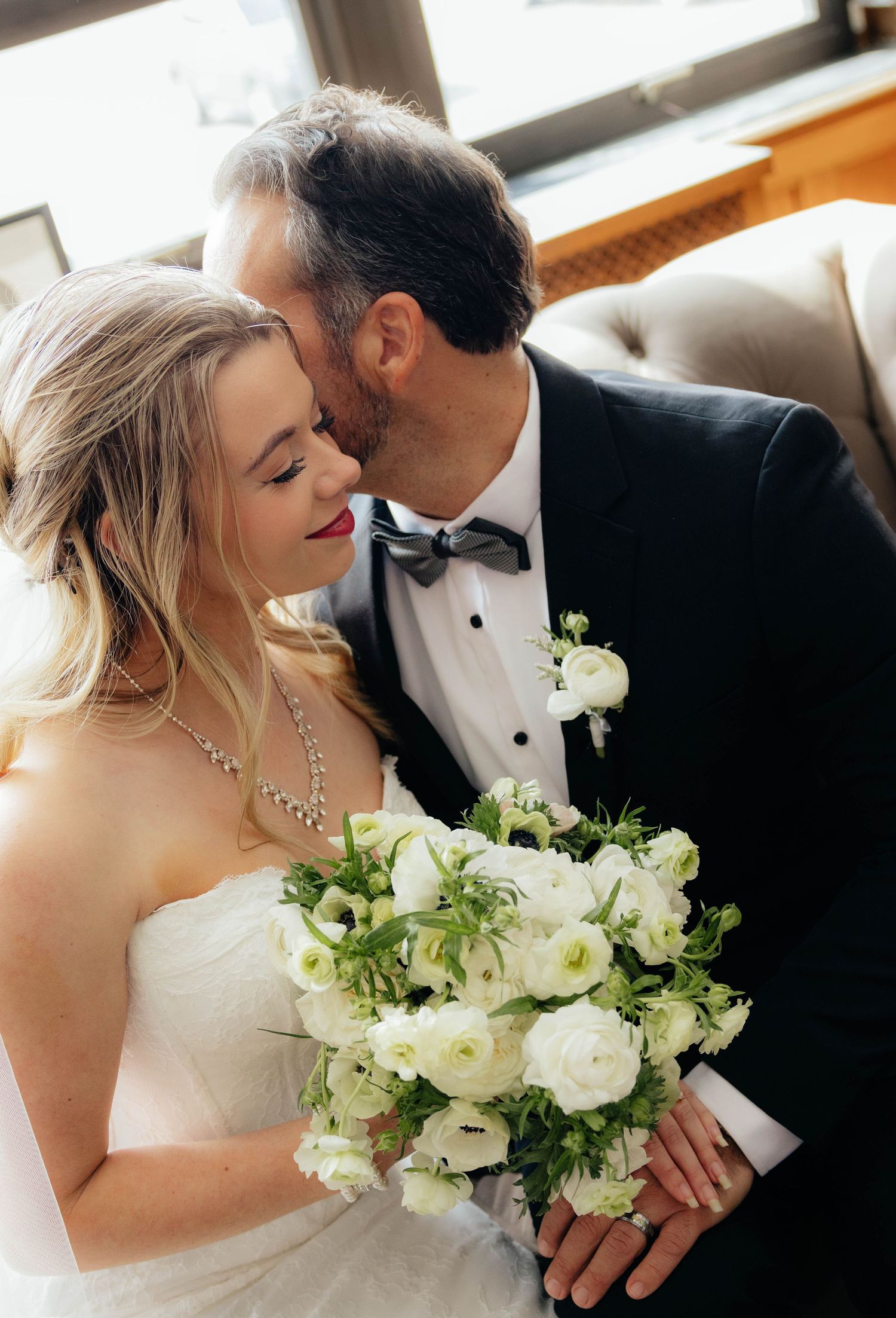 Groom in tuxedo kisses blonde bride holding white floral bouquet indoors.