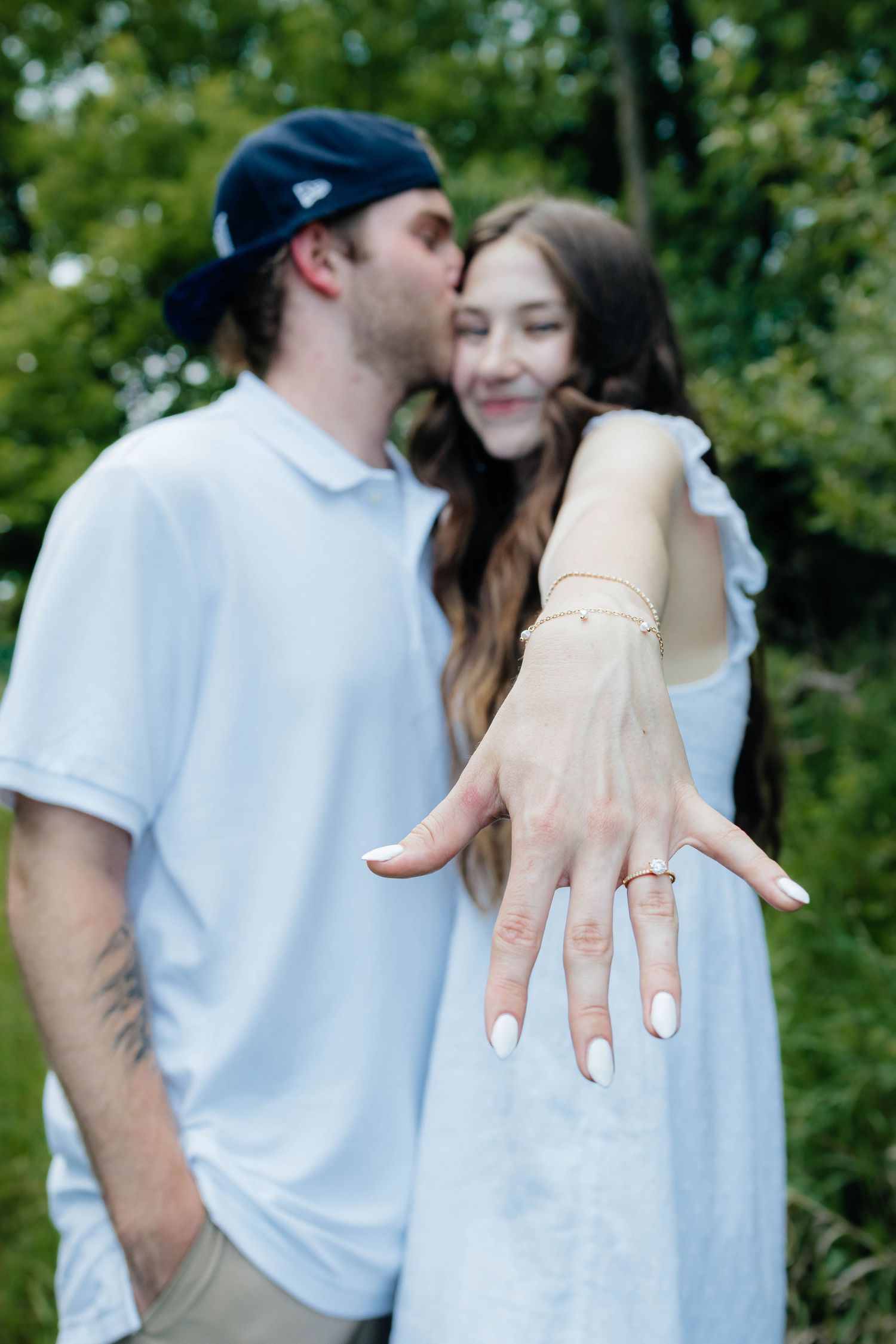 Couple showing off engagement ring as man kisses woman on cheek in green outdoor setting.
