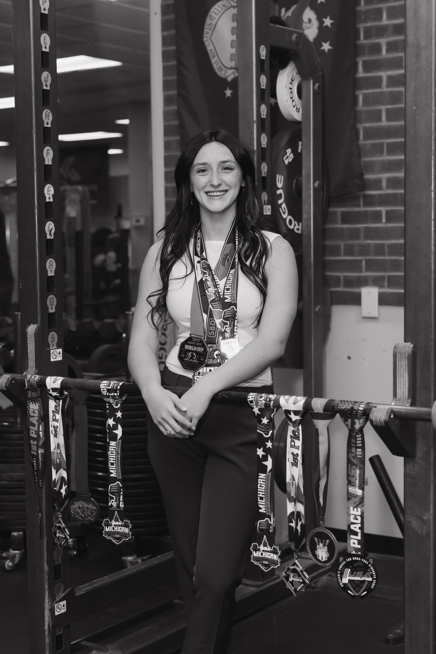 Black and white photo of a smiling woman displaying numerous athletic medals in a gym setting.