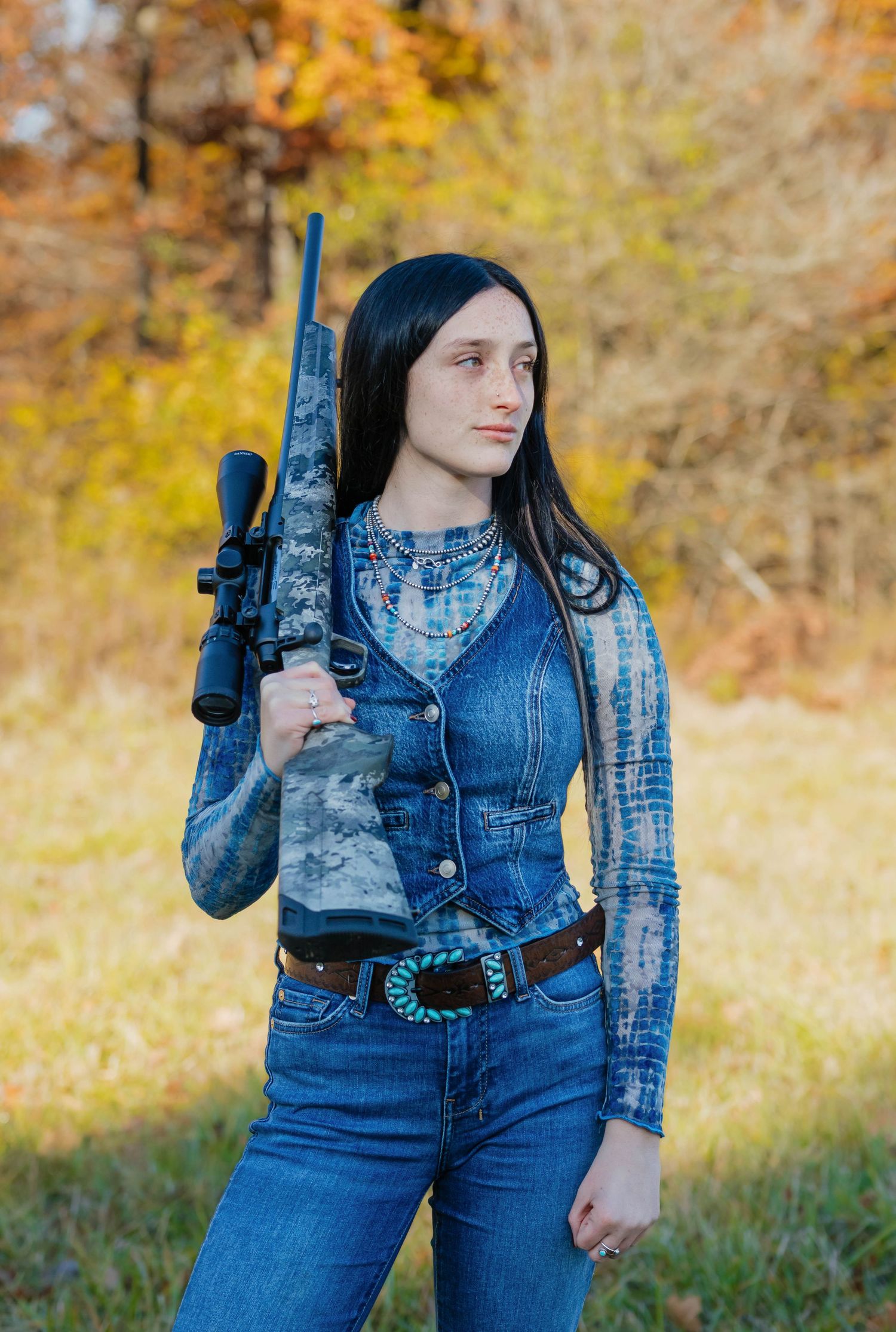Young woman in denim vest holding a camouflage hunting rifle in an autumn outdoor setting.