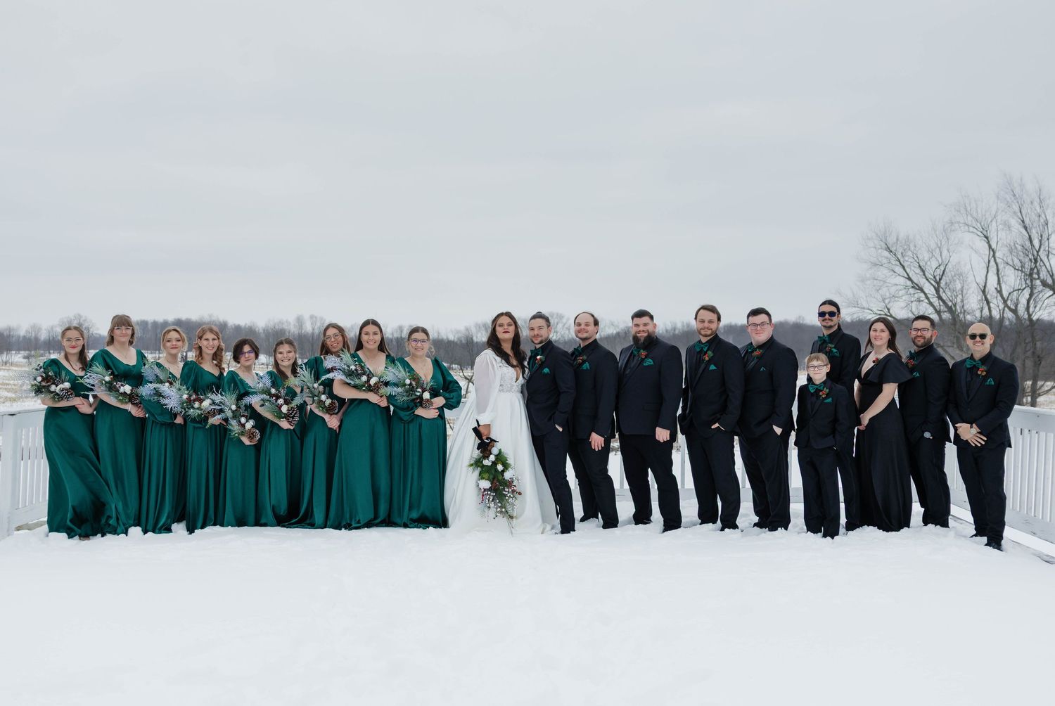 Large winter wedding party poses in snow, bridesmaids in emerald green gowns and groomsmen in black suits.