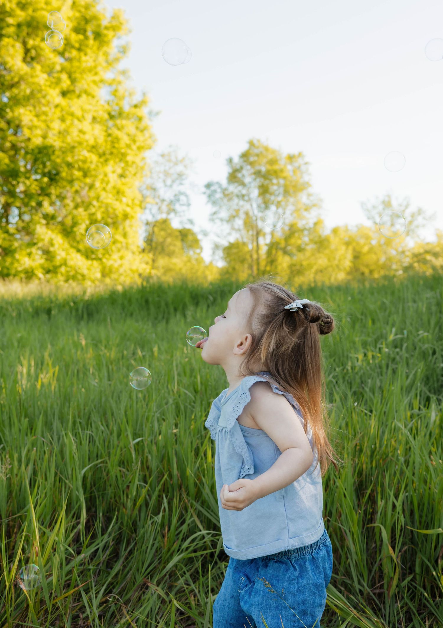 Young girl in a blue top looking up at soap bubbles floating in a sunny green meadow.