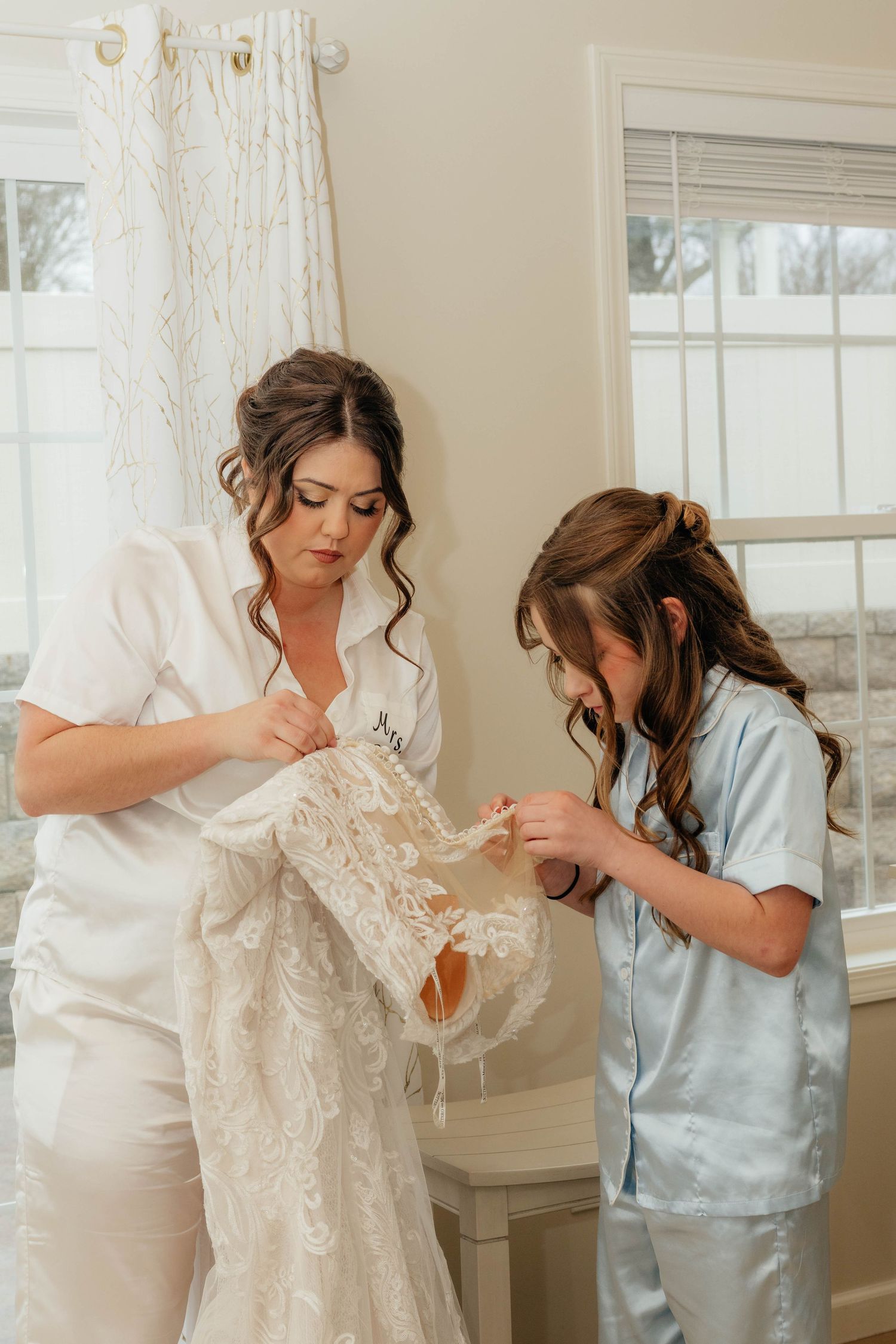 Bride in white robe examining lace wedding dress details with young girl in blue pajamas.