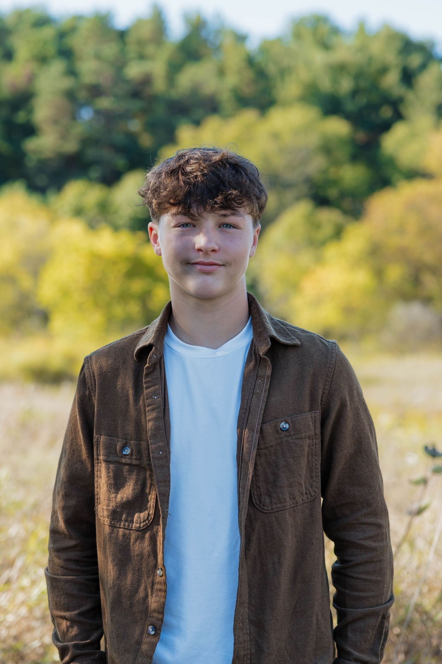 Smiling teenage boy with curly hair wearing a brown jacket in an autumn field.