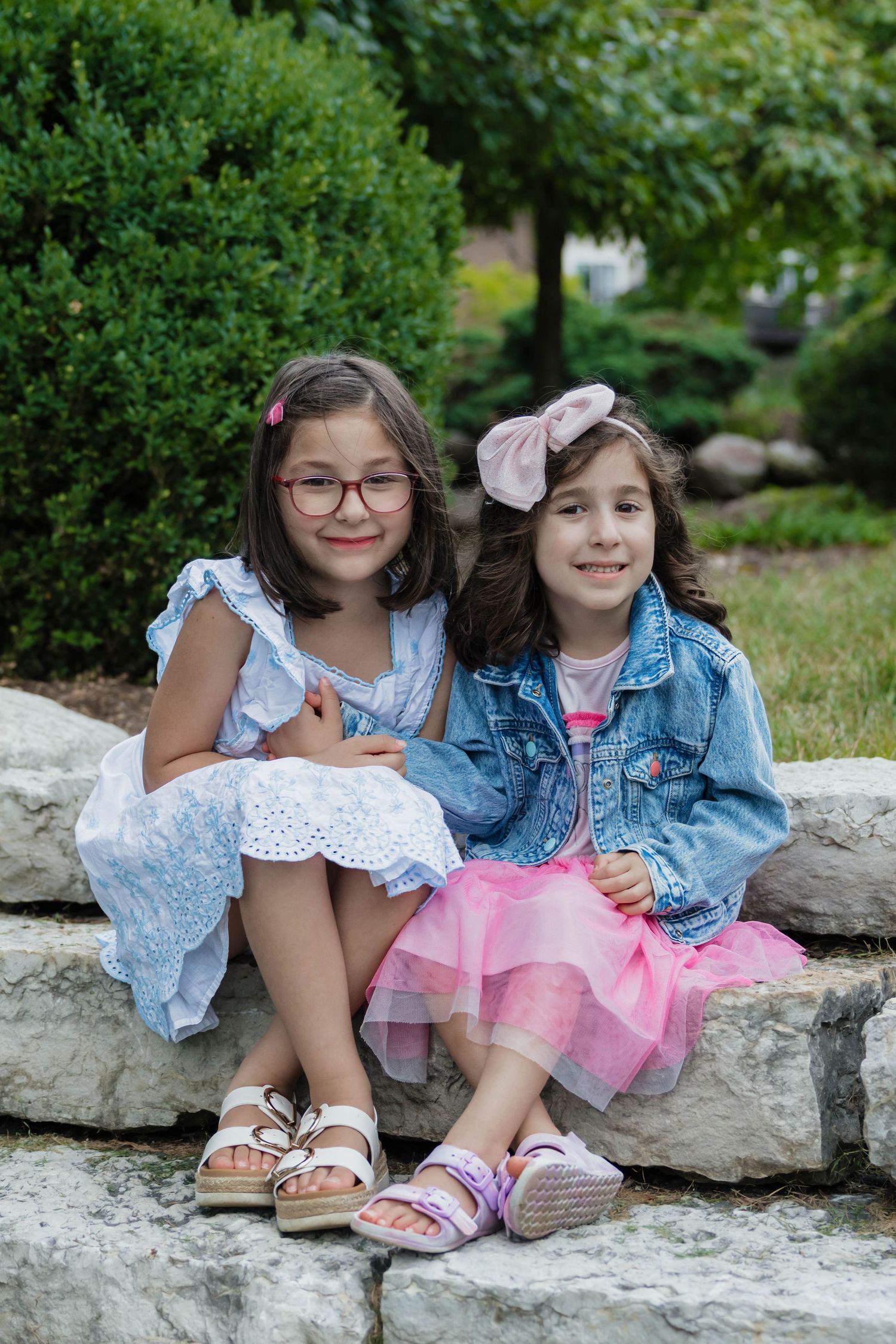 Two young girls sitting on stone steps in a garden, one wearing glasses and a blue dress, other in pink tutu.