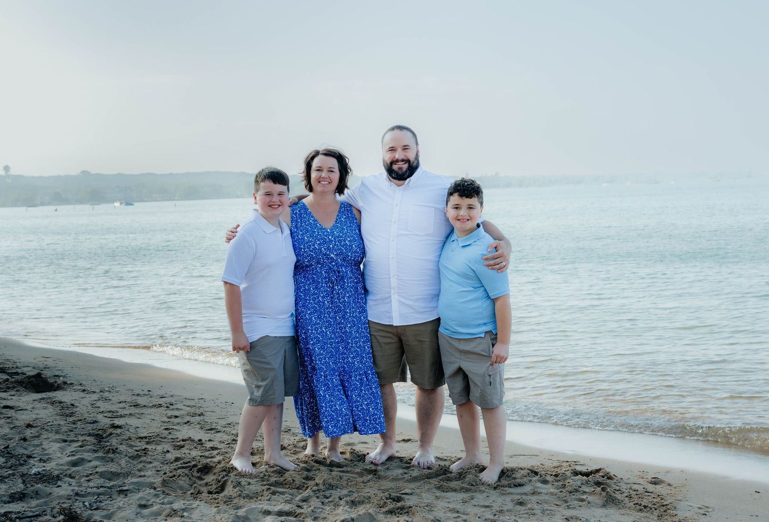 Family of four smiling together on a sandy beach with calm ocean water behind them.