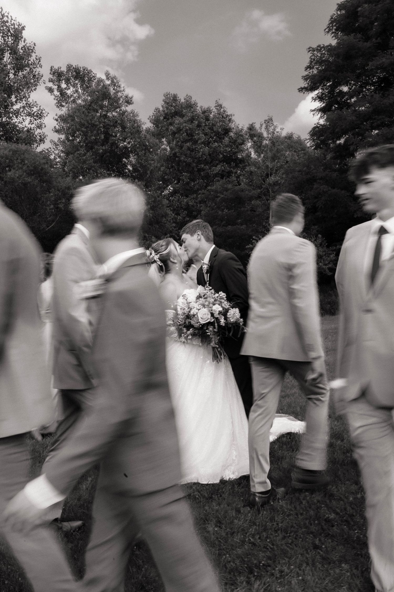 Black and white photo of bride and groom kissing while groomsmen walk past them outdoors at a wedding.