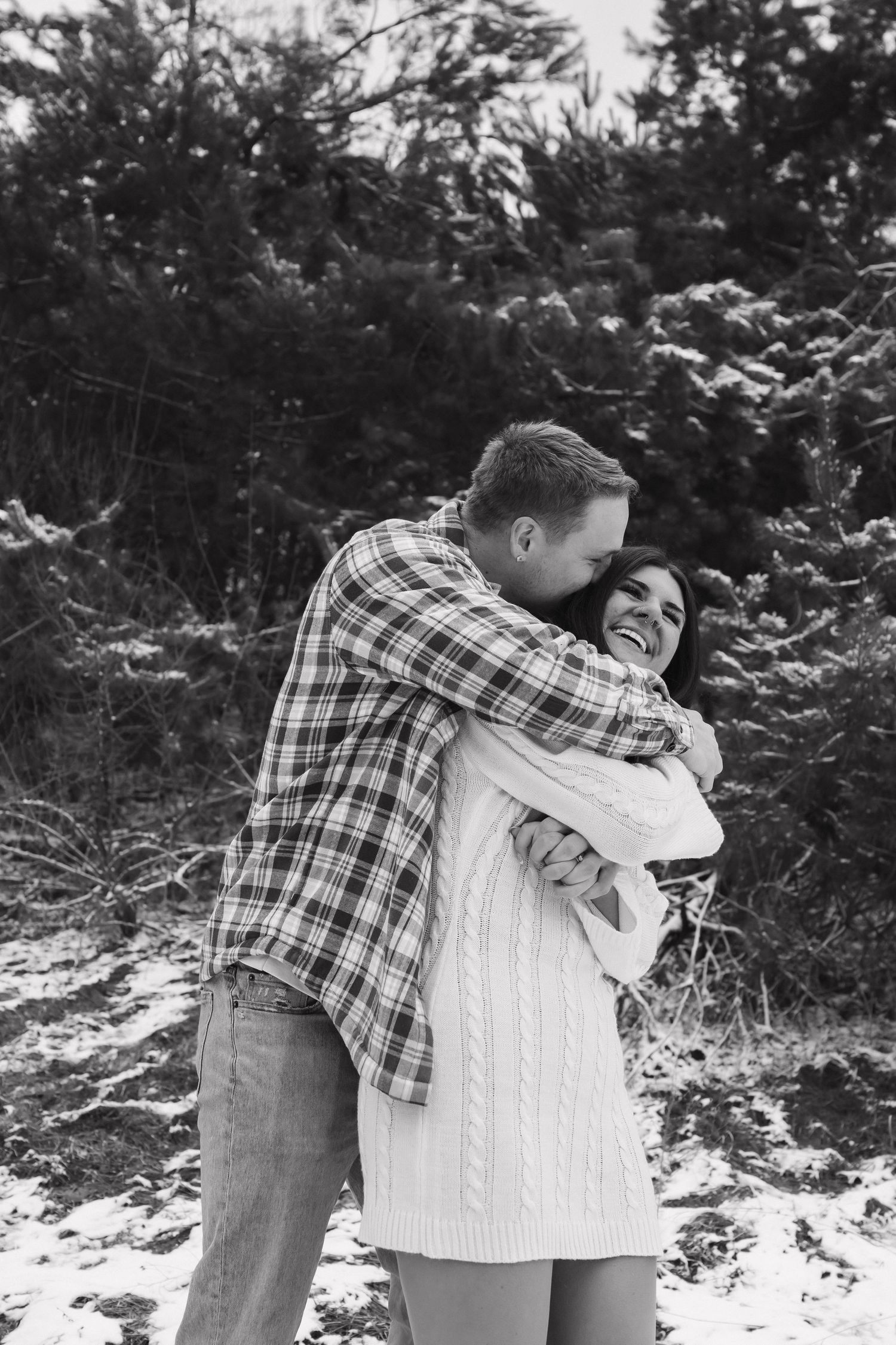 Black and white photo of a couple embracing and laughing together in a snowy outdoor winter setting.