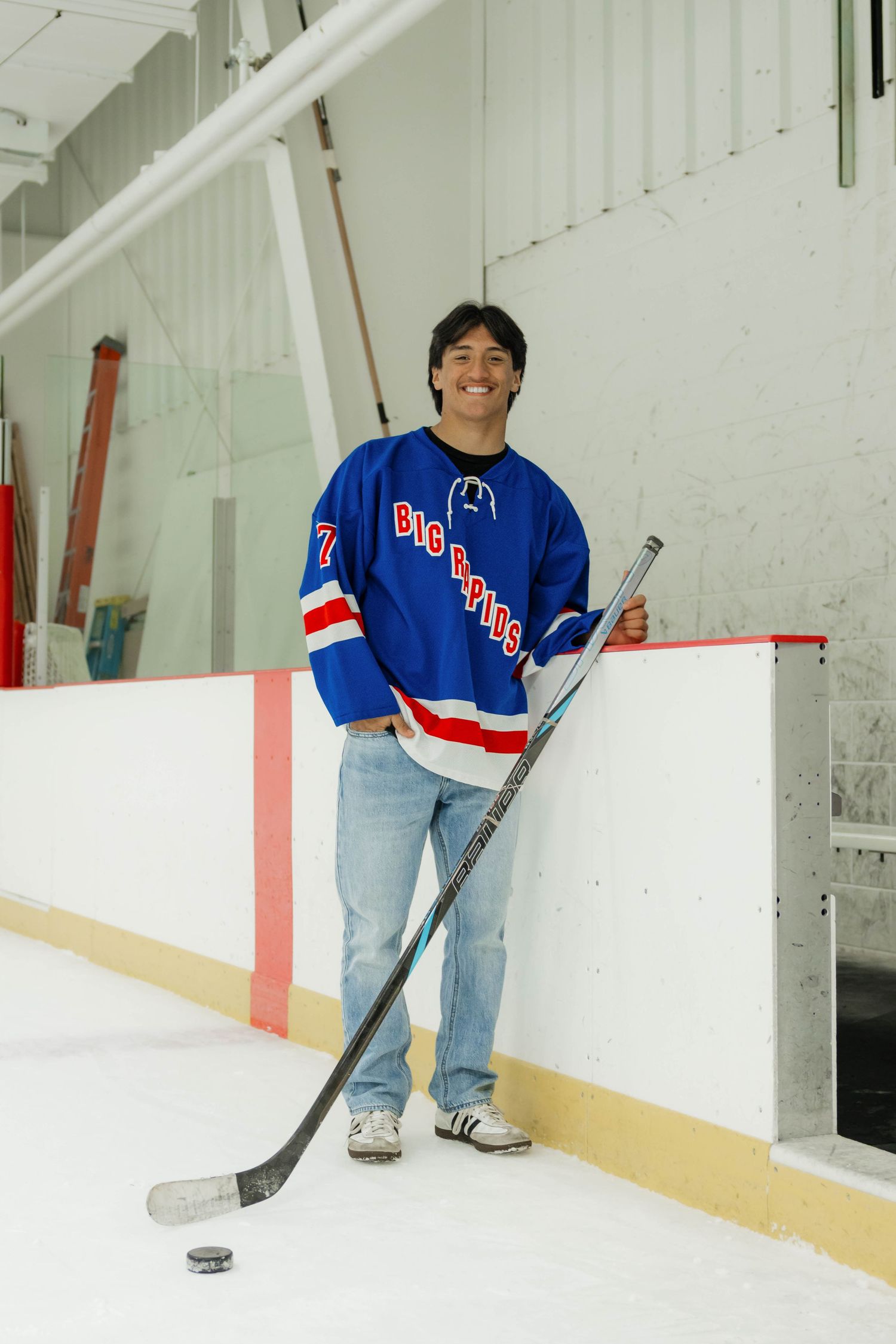 Teen in Big Rapids blue hockey jersey poses with stick and puck on ice rink.