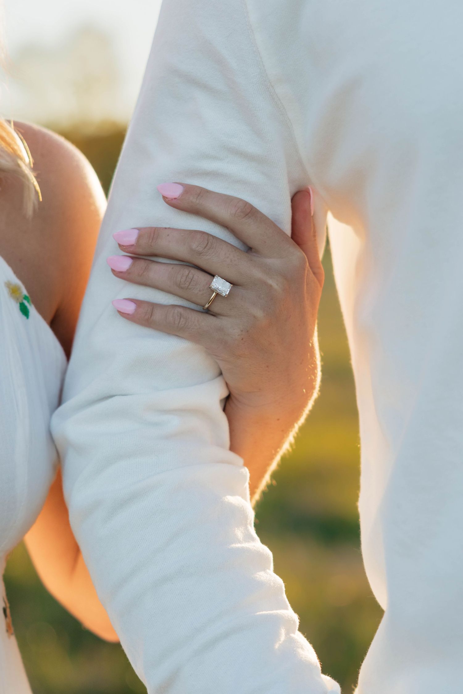 Close-up of a woman's hand with a diamond engagement ring resting on a man's arm outdoors at sunset.