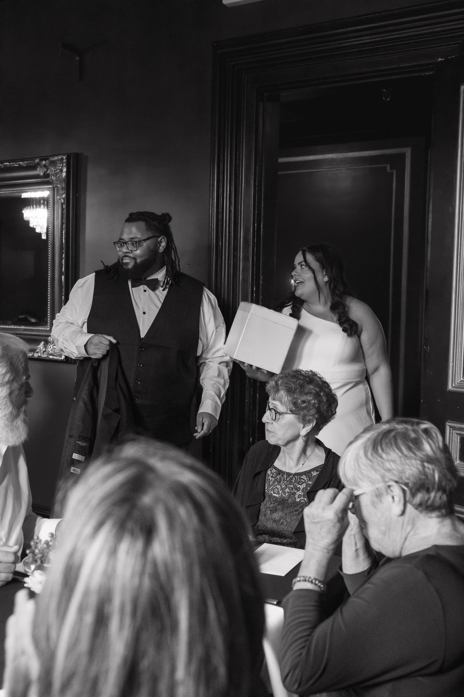 Bride and groom addressing wedding guests at elegant dark-walled reception venue, black and white photo.