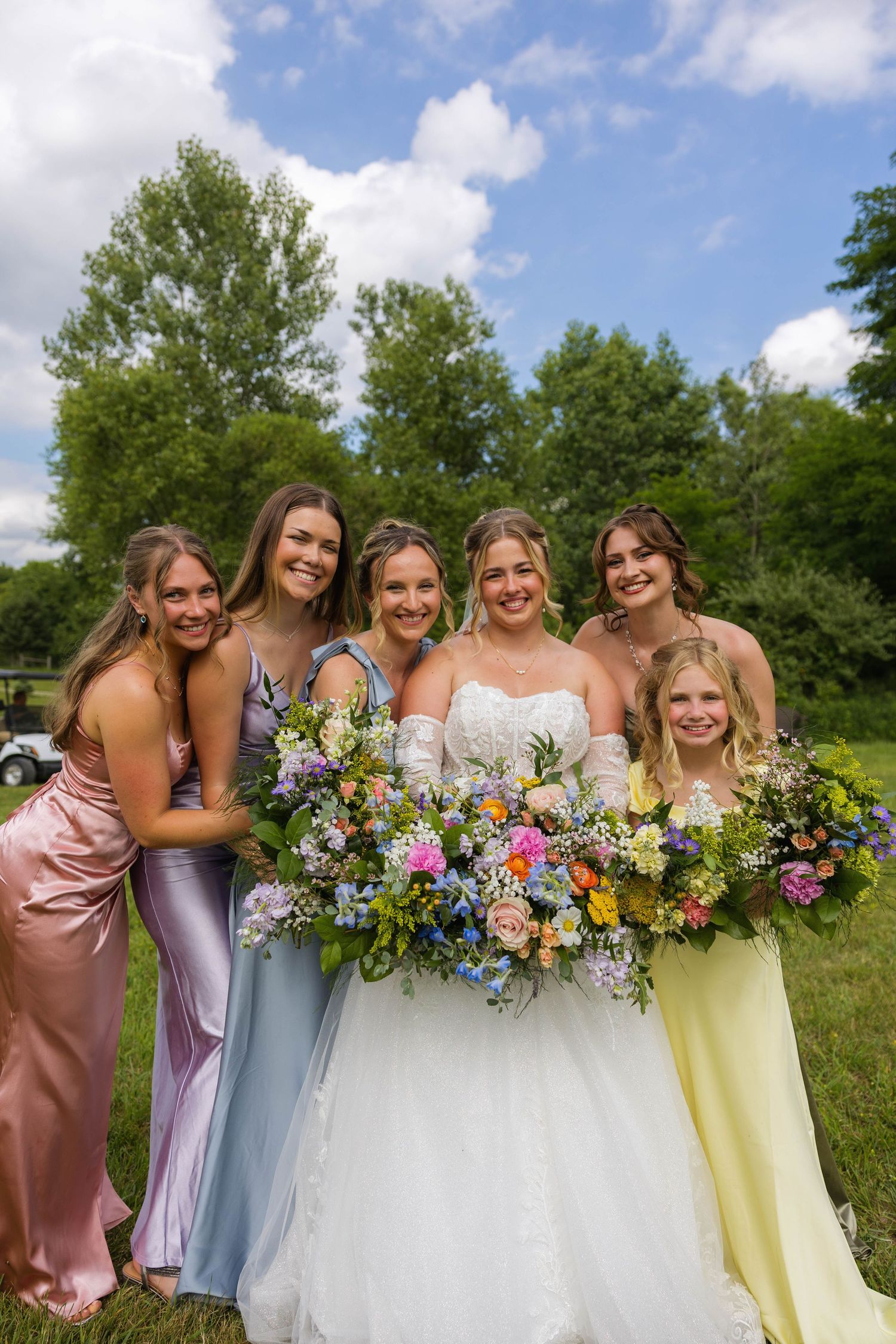 Bride in white gown with bridesmaids in colorful dresses holding wildflower bouquets outdoors on a sunny day.