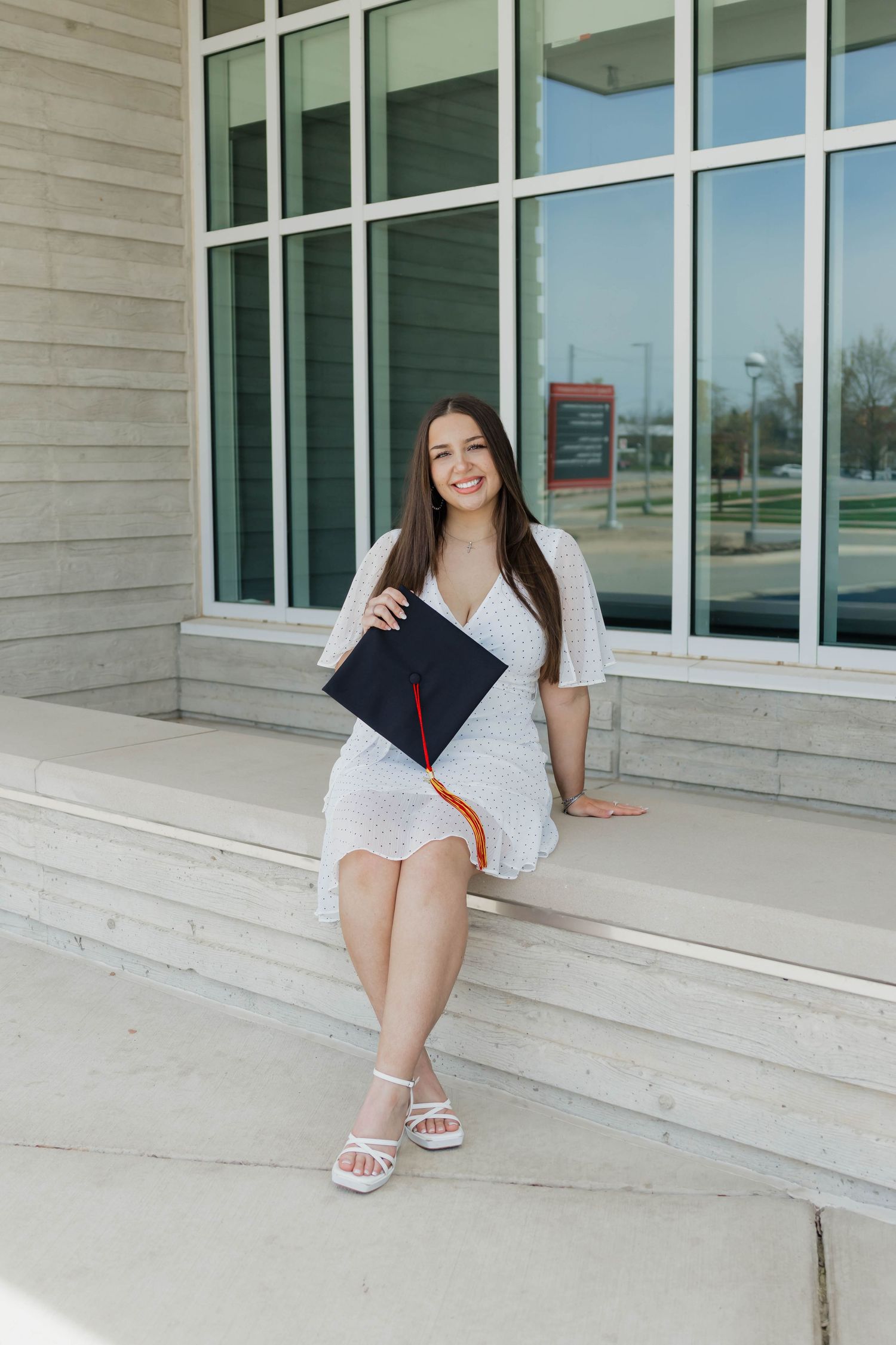 Young woman in white dress holding graduation cap sits on steps outside modern building, celebrating academic achievement.